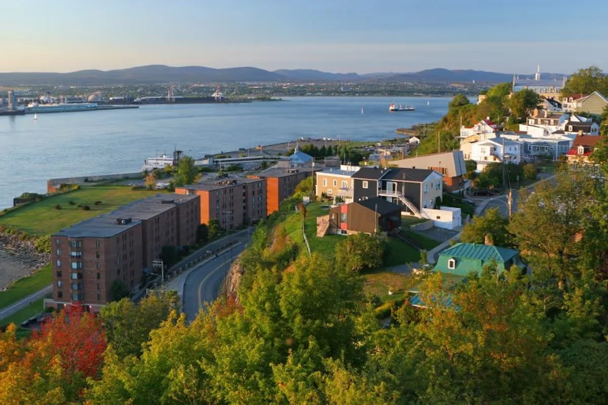 Vues d'une ville côtière avec des maisons colorées, des bâtiments résidentiels, un port avec des navires, et des montagnes à l'arrière-plan.