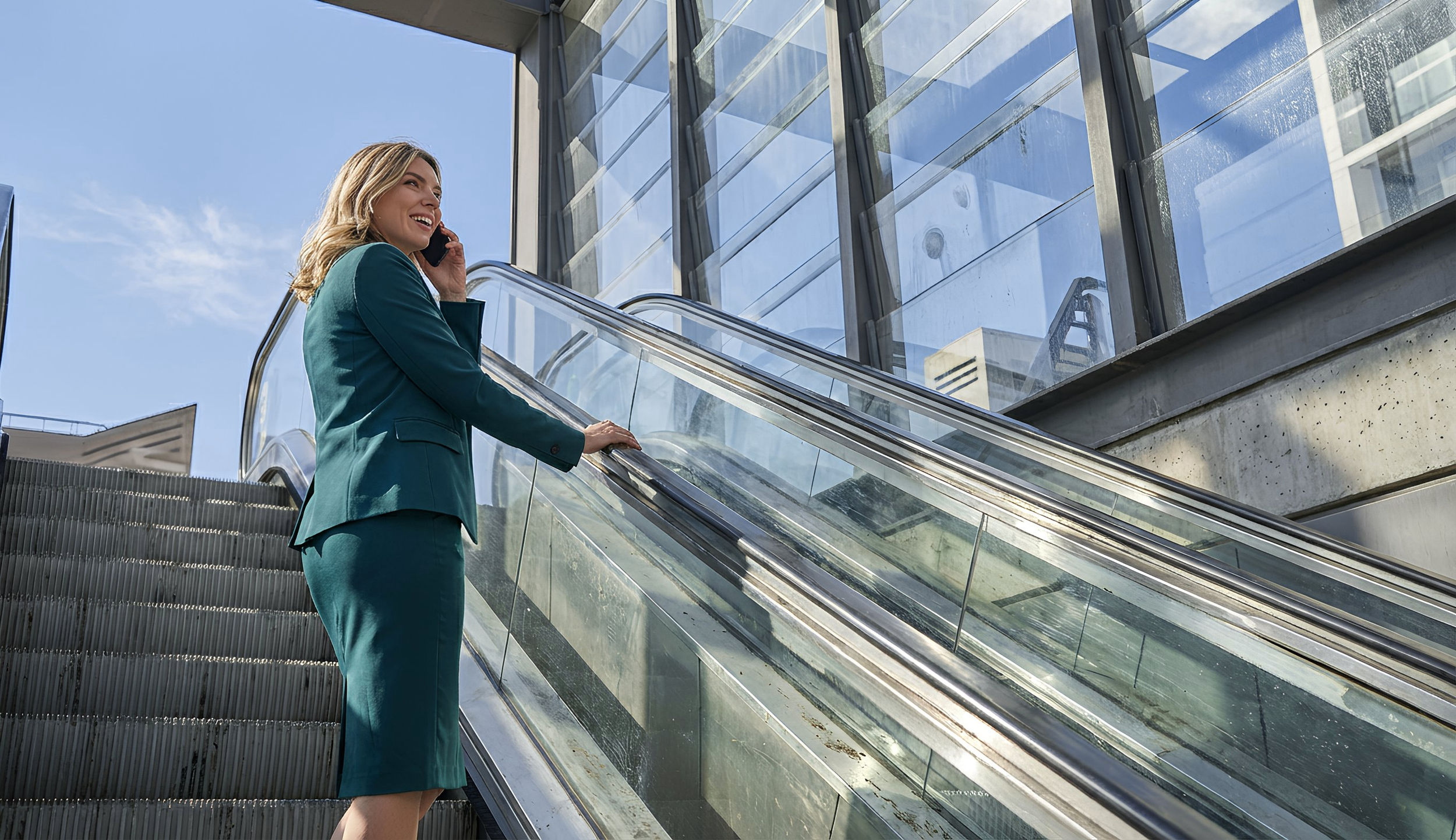 Femme d'affaires parlant au téléphone tout en montant un escalator devant un bâtiment moderne en verre.