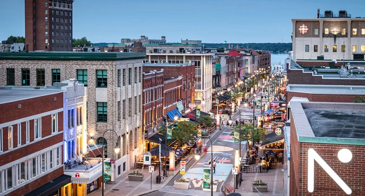 Rue commerçante animée en soirée avec boutiques, bars, et restaurants, bâtiments en briques, lampposts, et vue sur le fleuve en arrière-plan.