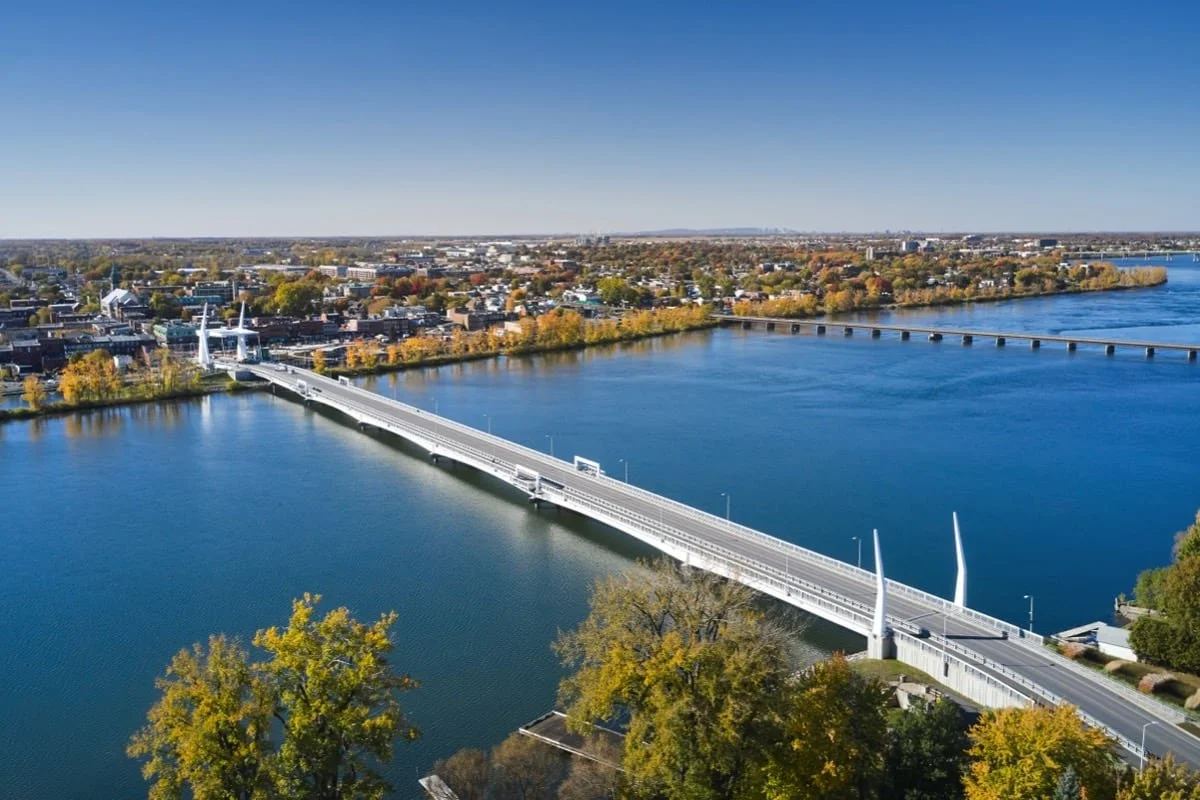 Vue aérienne d'un pont moderne traversant une rivière, avec une ville en arrière-plan et des arbres aux couleurs automnales.