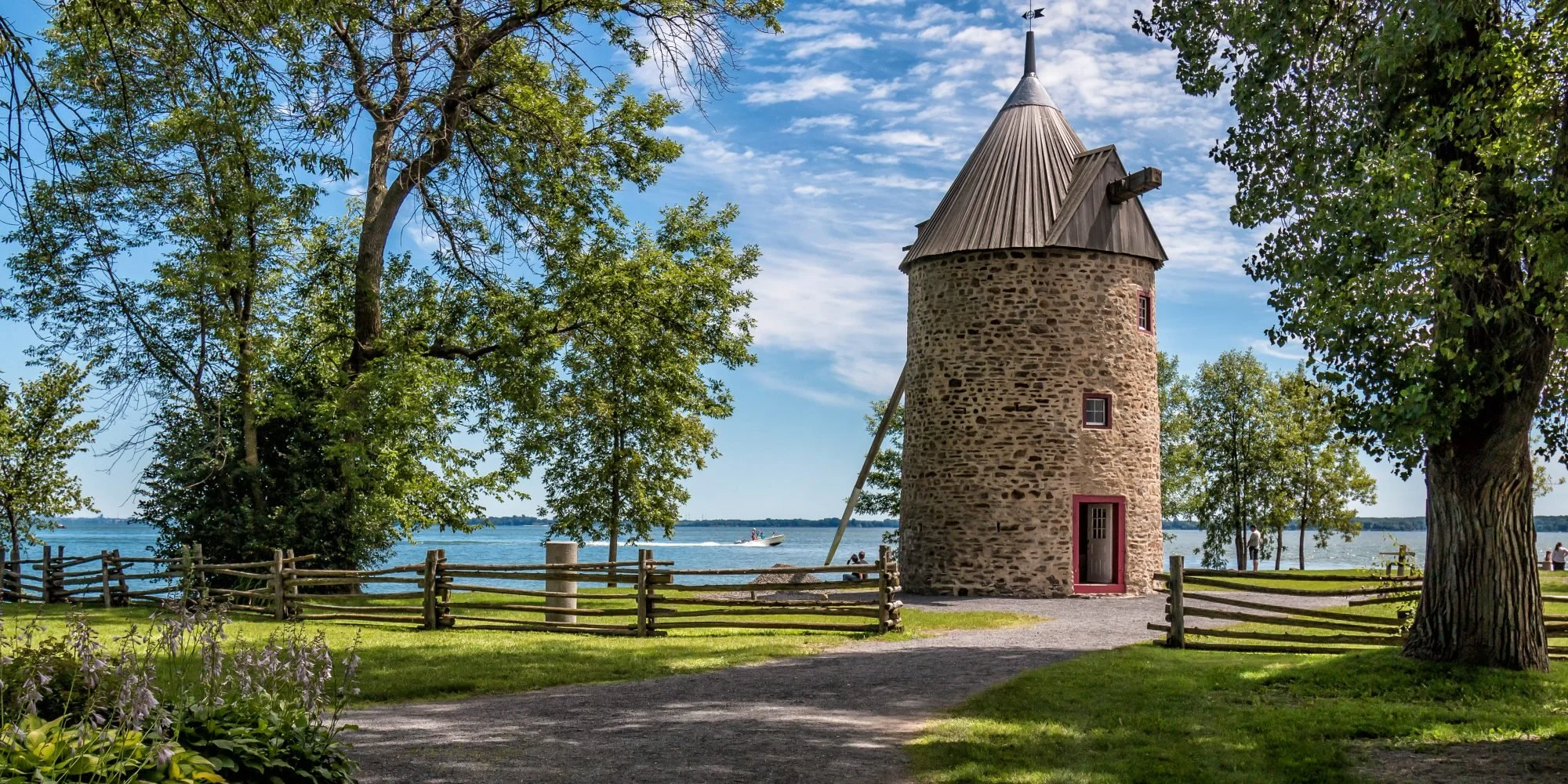 Tour en pierre ronde avec toiture en forme de cône, situé près d'un lac, entouré d'arbres verts et d'une clôture en bois, avec un bateau à moteur sur l'eau et des personnes à proximité.