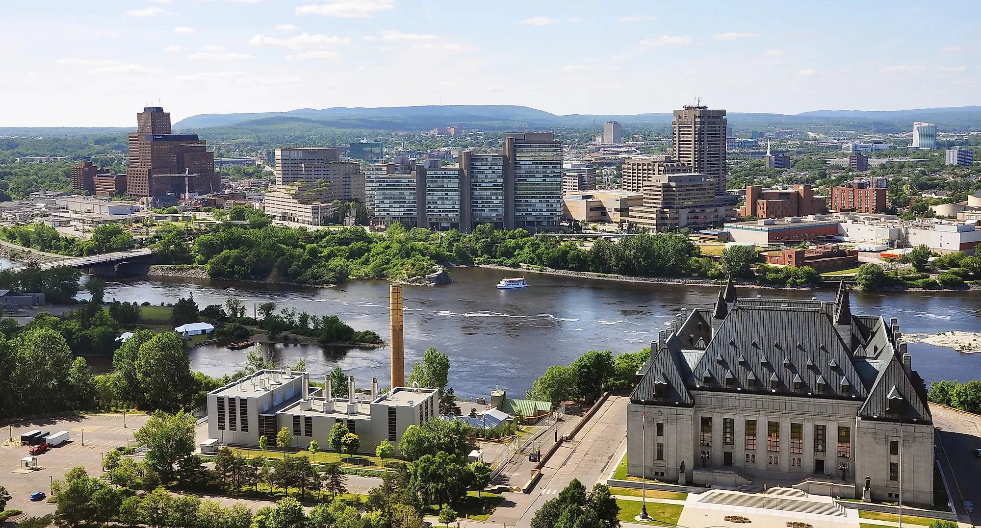 Vue panoramique d'une ville avec un fleuve au premier plan, des bâtiments modernes et un paysage de montagnes en arrière-plan, par une journée ensoleillée.