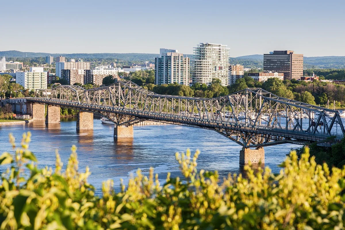 Un pont métallique traverse une rivière avec des bâtiments modernes en arrière-plan, sous un ciel clair.