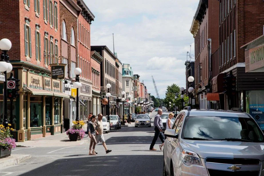 Rue animée avec des bâtiments en brique, des personnes marchant, des voitures garées, lampadaires avec lampes blanches, fleurs et commerce locaux.