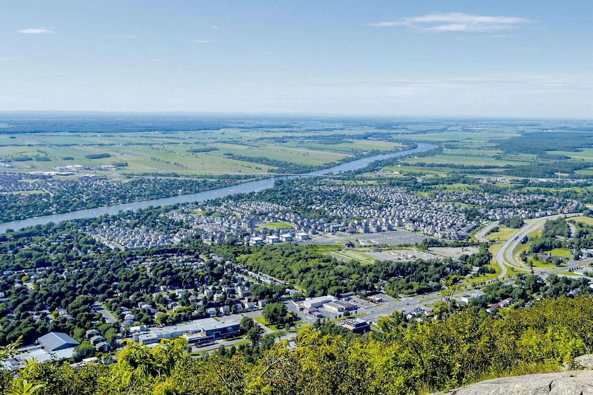 Vue aérienne d'une ville avec rivière, maisons, zones vertes, et routes sous un ciel clair.