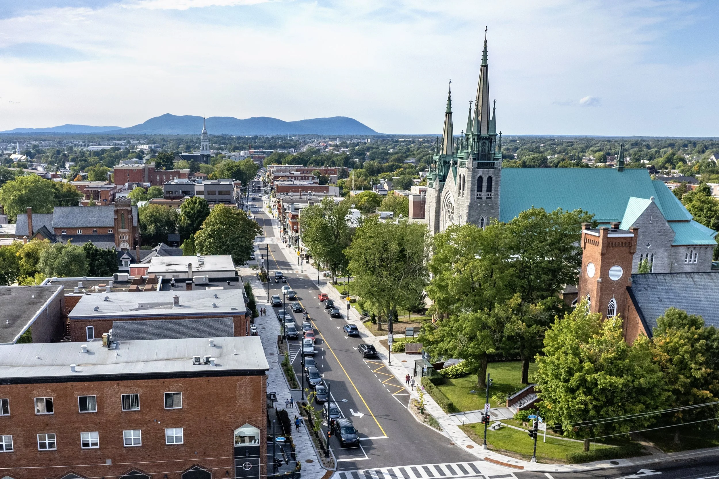 Une vue aérienne d'une ville avec une église gothique, des bâtiments résidentiels et commerciaux, des rues avec des voitures, de nombreux arbres, et des montagnes à l'horizon.