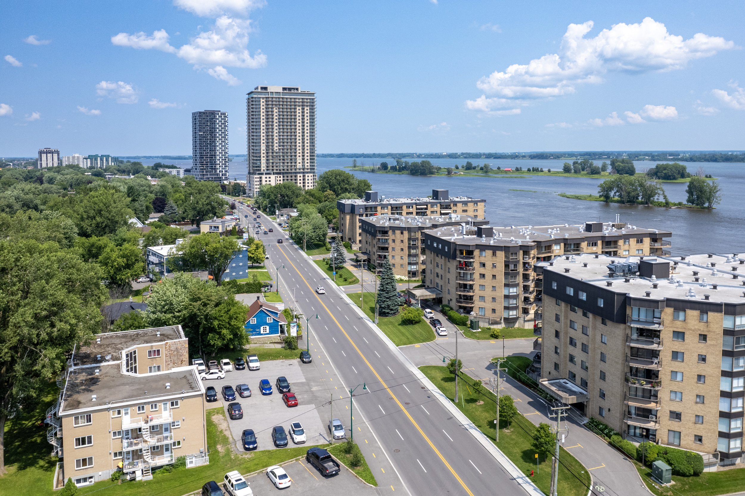 Vue aérienne d'une ville avec des immeubles résidentiels, une route passante, une zone boisée, et un fleuve en arrière-plan, sous un ciel bleu avec quelques nuages.