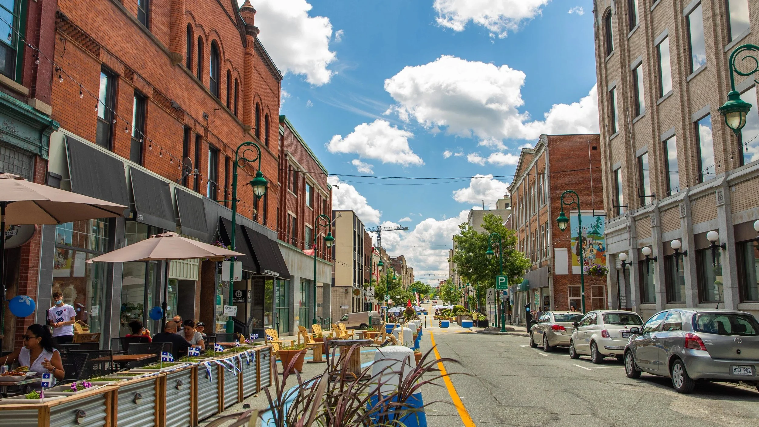 Rue commerçante animée avec des bâtiments en briques rouges et beige, des enseignes de magasins, des parasols beige pour une terrasse de café ou restaurant, personnes assises, ciel bleu avec des nuages, arbres, voitures garées et circulation, lampadaires verts.