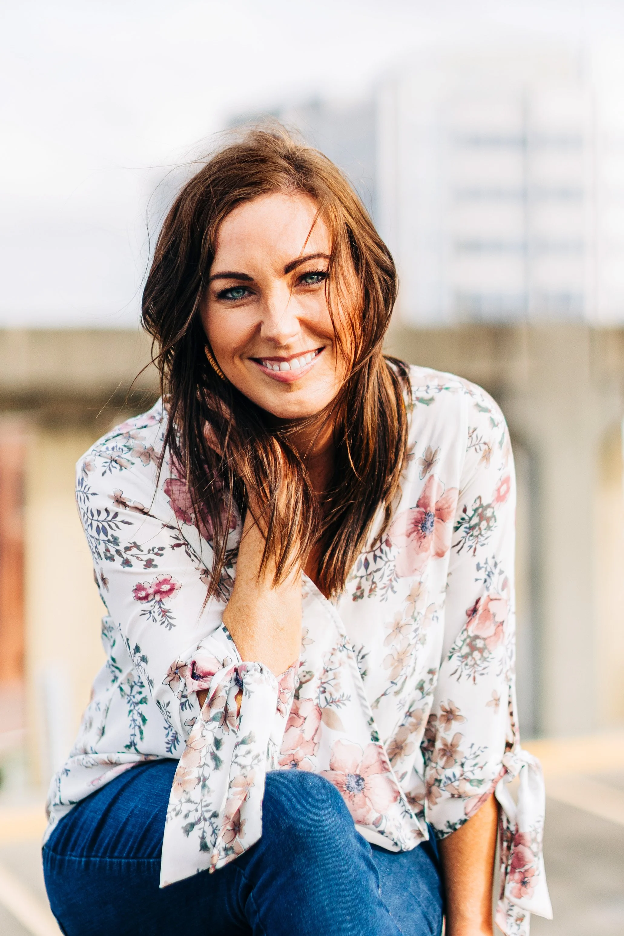 A smiling woman with long brown hair and blue eyes, wearing a floral blouse and blue jeans, sitting outdoors with a blurred cityscape background.