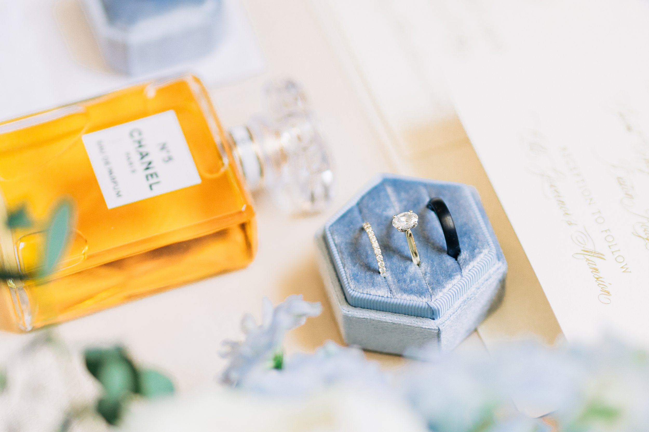 A close-up photo of jewelry including rings and a bracelet displayed in a blue velvet box, with perfume bottles and a handwritten note in the background.