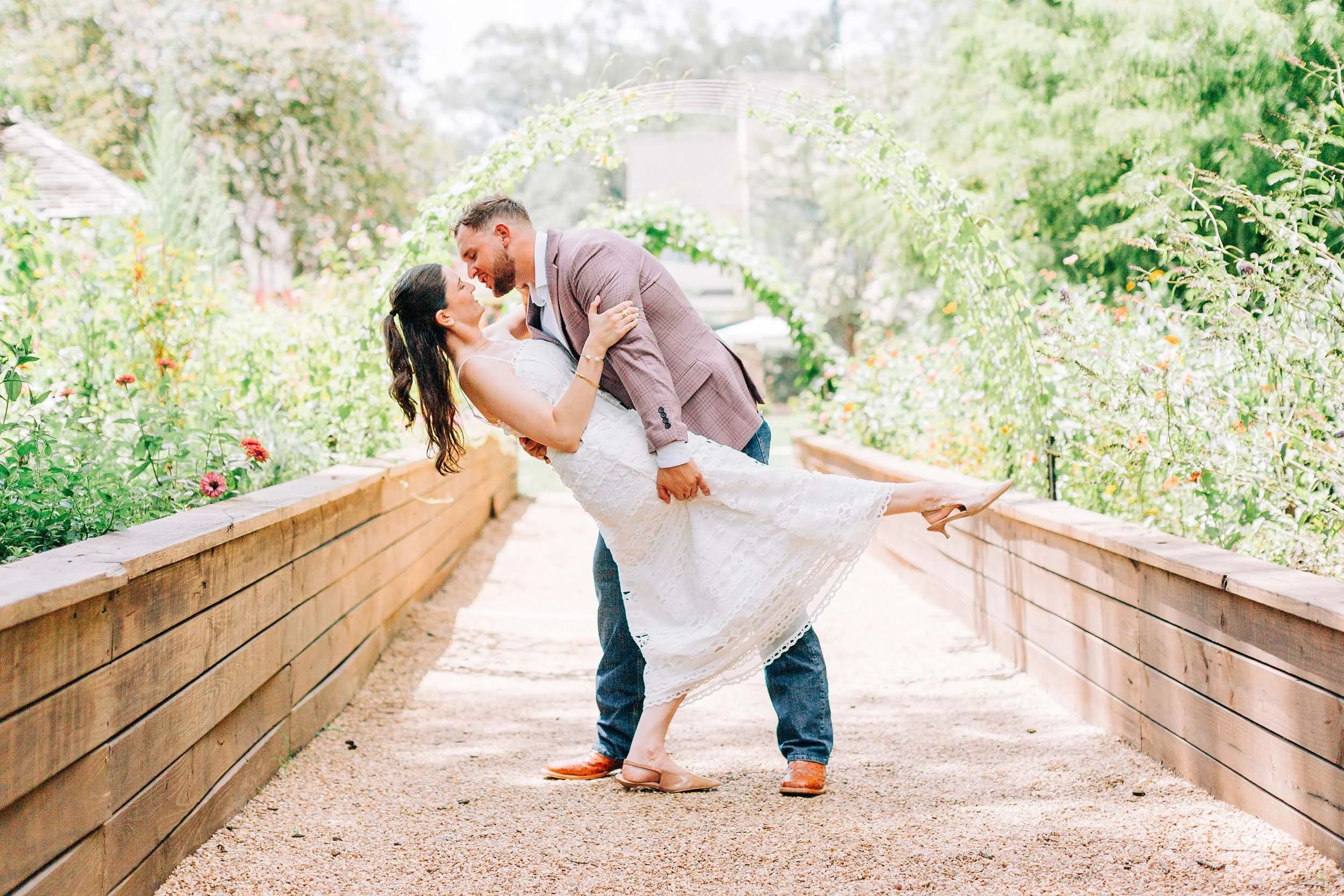 A couple dancing in a garden with lush green trees and a wooden path. The man is wearing a checkered blazer and jeans, and the woman in a white lace dress. The man is holding the woman in a dip, and they are smiling at each other.