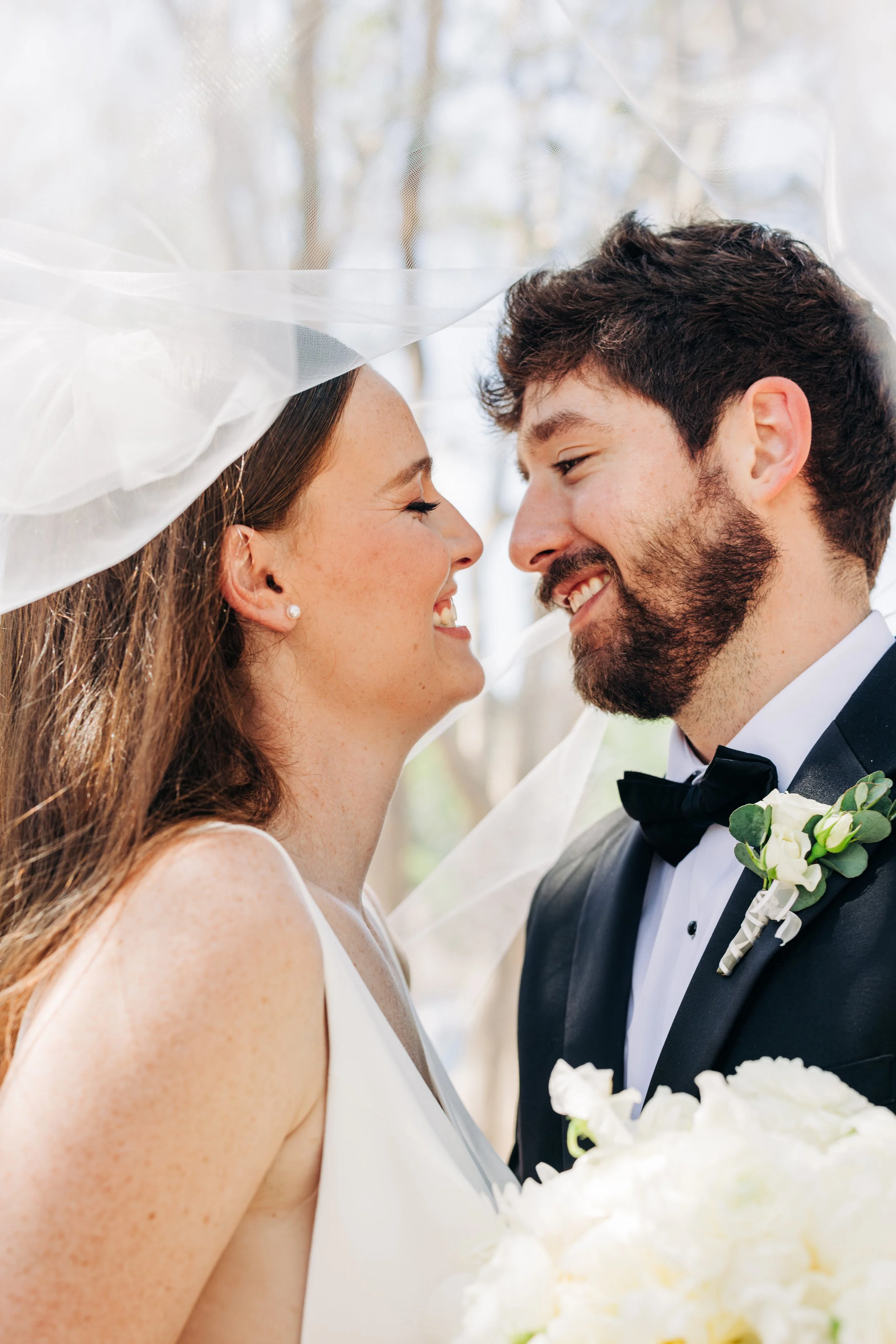 Close-up of a bride and groom smiling closely during wedding, with wedding veil visible on bride and groom wearing tuxedo with boutonniere.