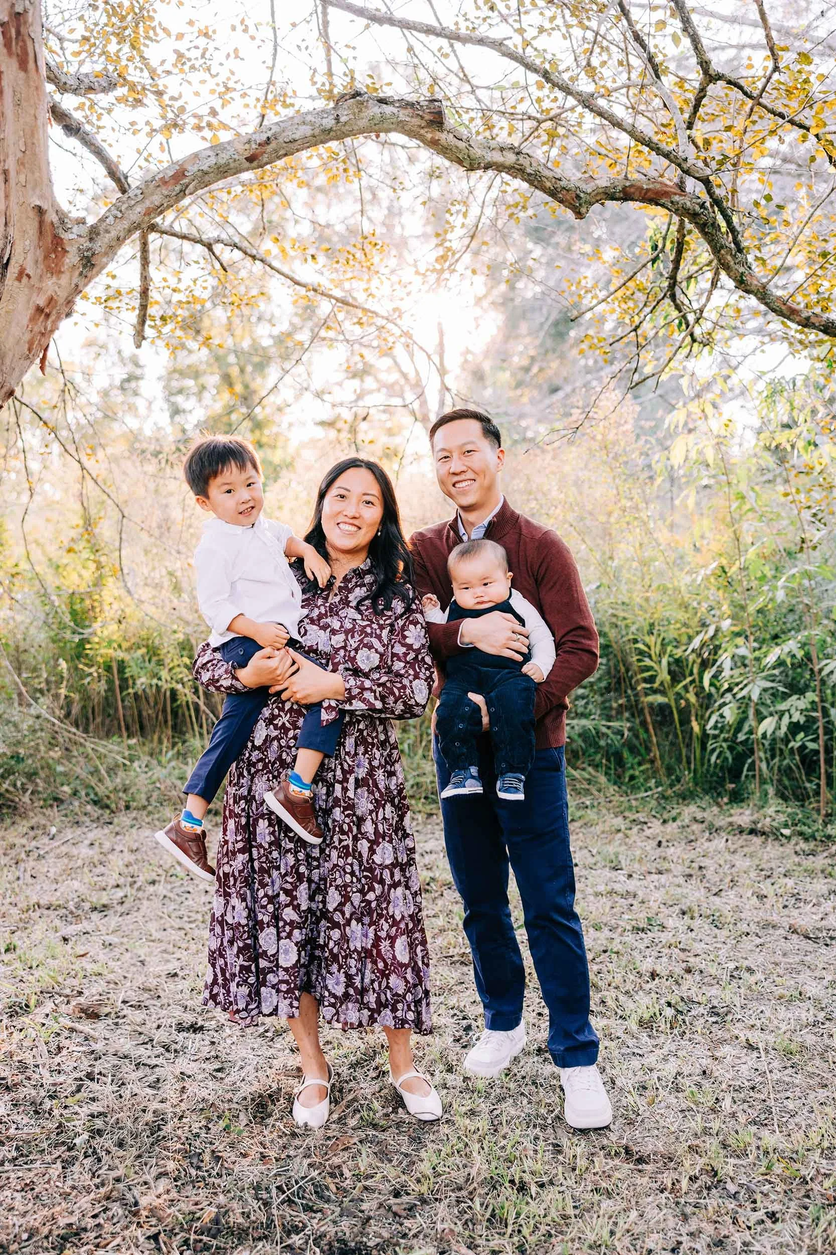 A family of four outdoors in a wooded area with fall foliage, smiling and posing for a photo. The mother is holding a young boy, and the father is holding a baby boy.