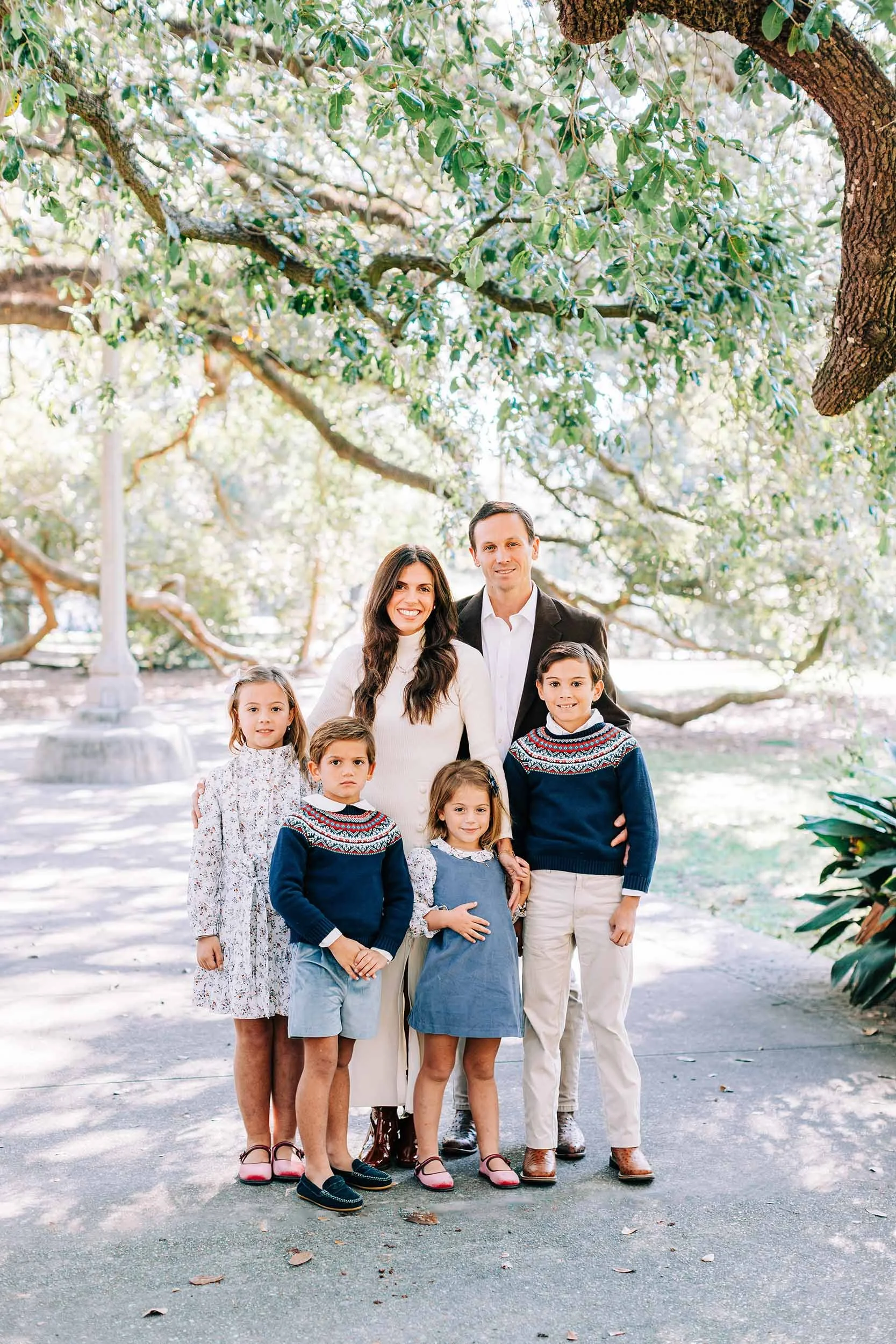 A family group photo outdoors in a park with trees in the background. The family consists of two adults and five children standing together, smiling at the camera.