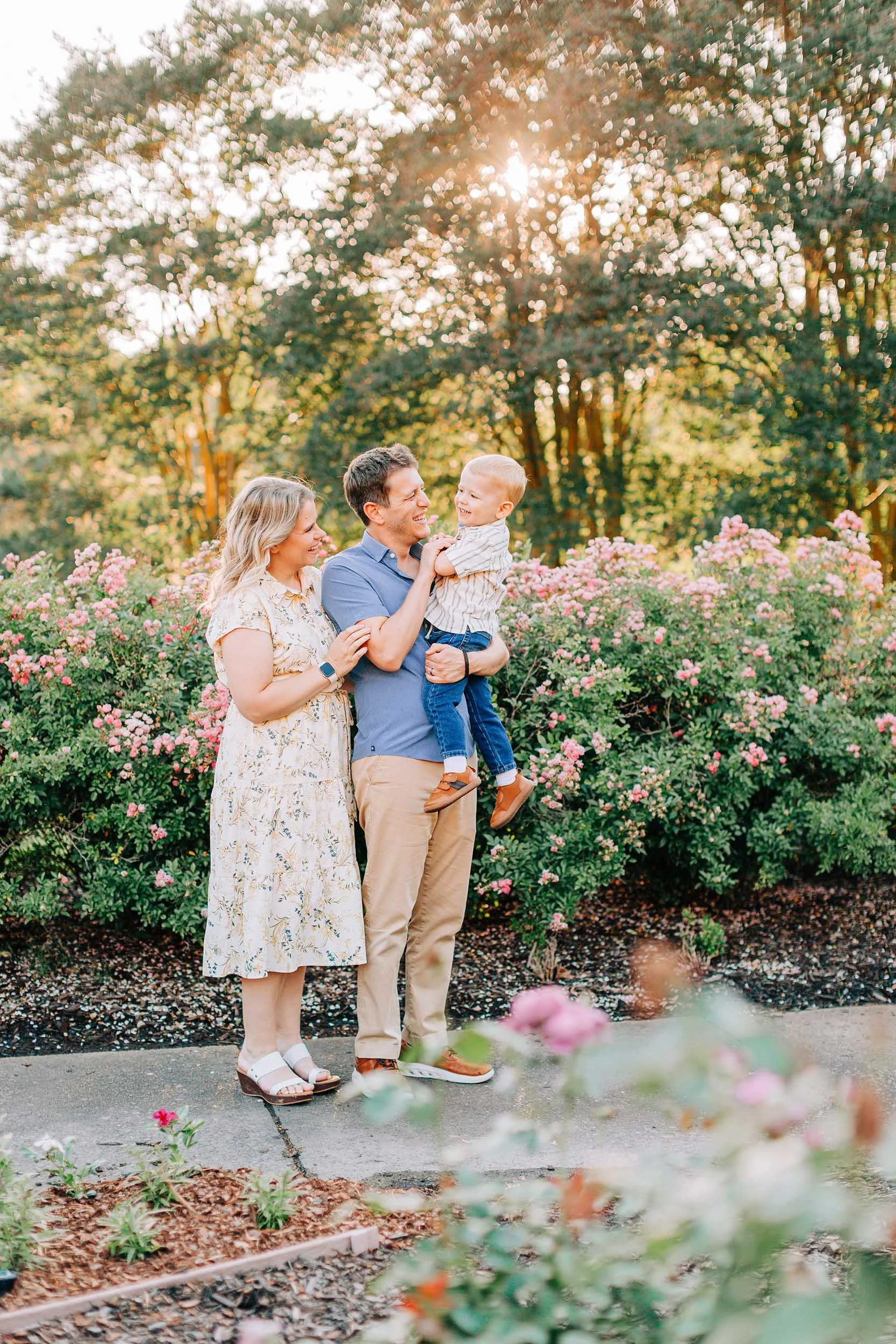 A family of three, a woman, a man, and a young boy, standing outdoors near pink bushes and flowers during golden hour sunlight. The man holds the boy and they are both smiling at each other while the woman looks on affectionately.