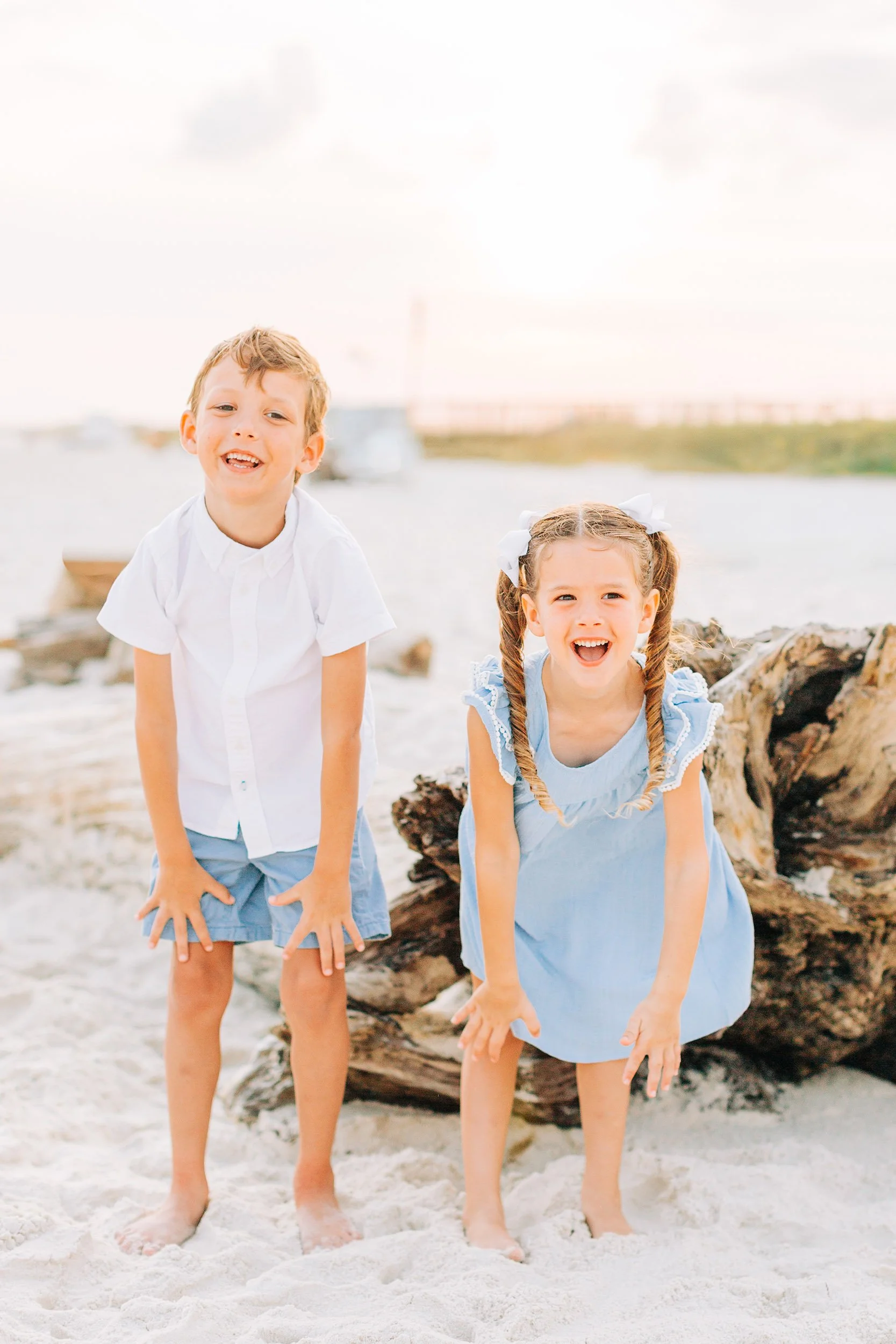 Two young children, a boy and a girl, playing on the beach in front of a large piece of driftwood, with water and a hazy sky in the background. They are smiling and appear to be happy.