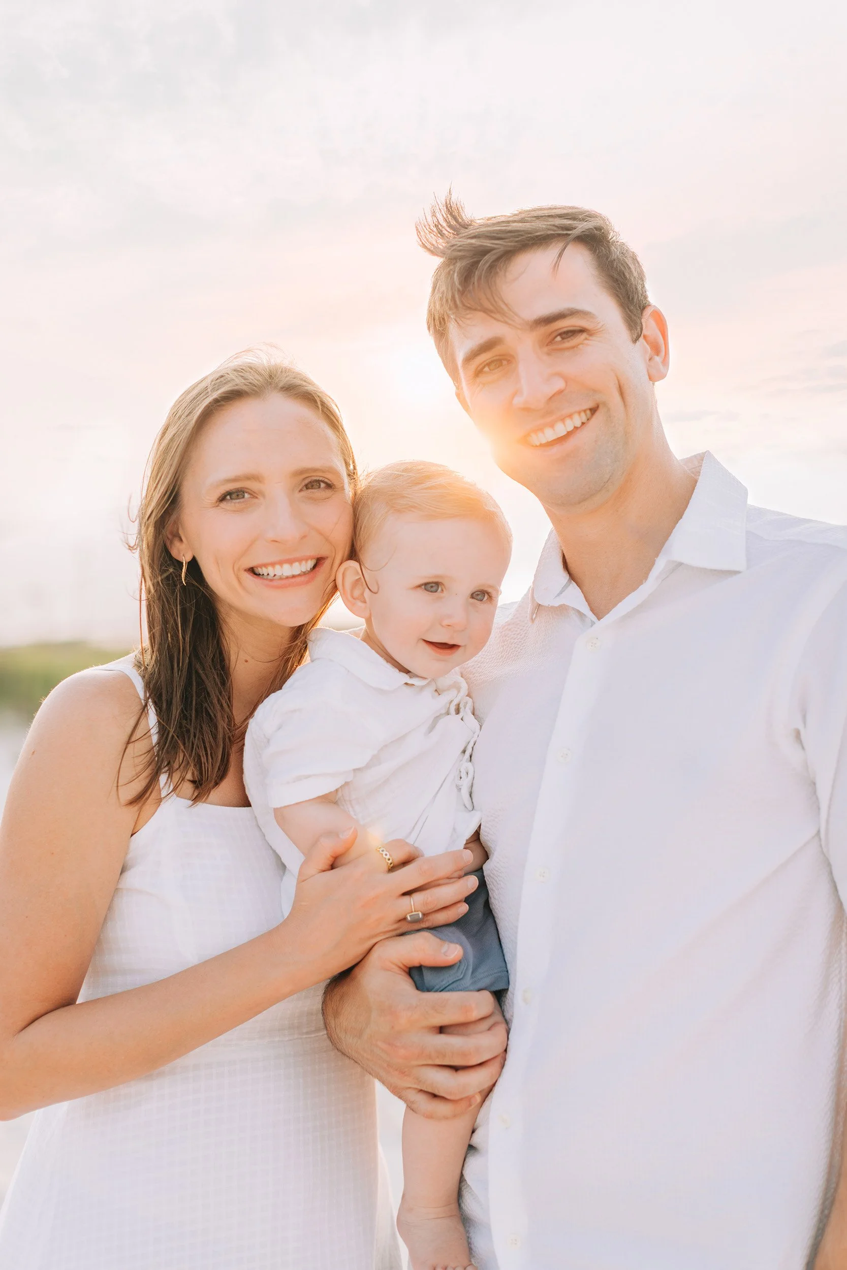 A happy family of three, a woman, a man, and a young child, smiling together outdoors during sunset. The woman has long brown hair, the man has short light brown hair, and the child has curly red hair. They are dressed in light-colored clothing.