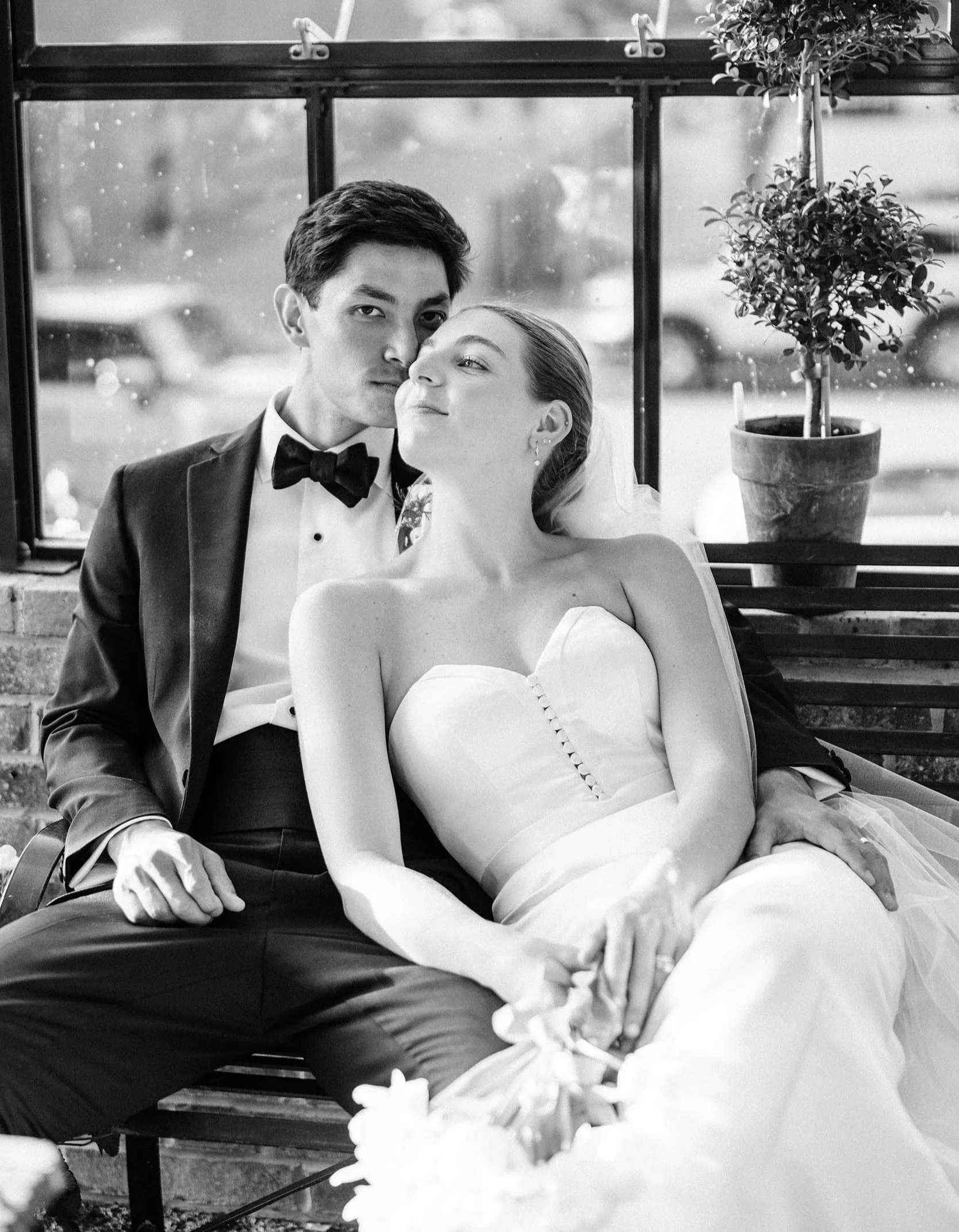 A black-and-white photo of a man and woman in wedding attire sitting on a bench indoors. The man is wearing a tuxedo with a bow tie, and the woman is in a strapless wedding gown with a smiling expression. They are close to each other with the woman's head tilted towards the man. There is a potted plant and raindrops on the window behind them.