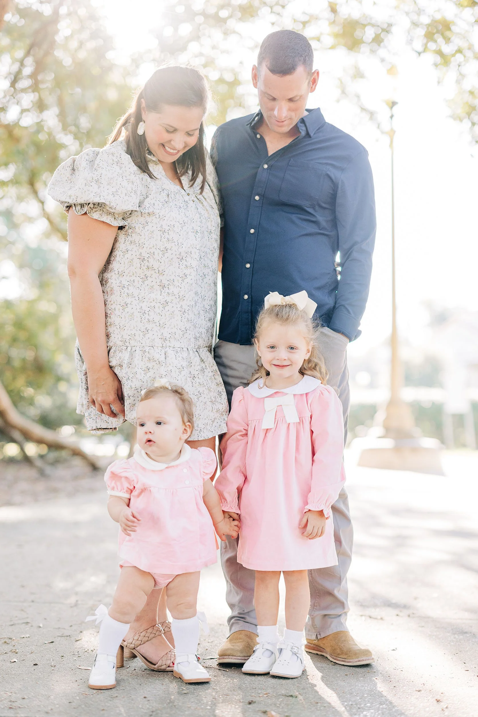 A family of four, including a mother, father, and two young daughters, standing outdoors on a sunny day. The mother is wearing a light-colored dress with puffed sleeves, and the father is dressed in a navy blue shirt and beige pants. The daughters ar