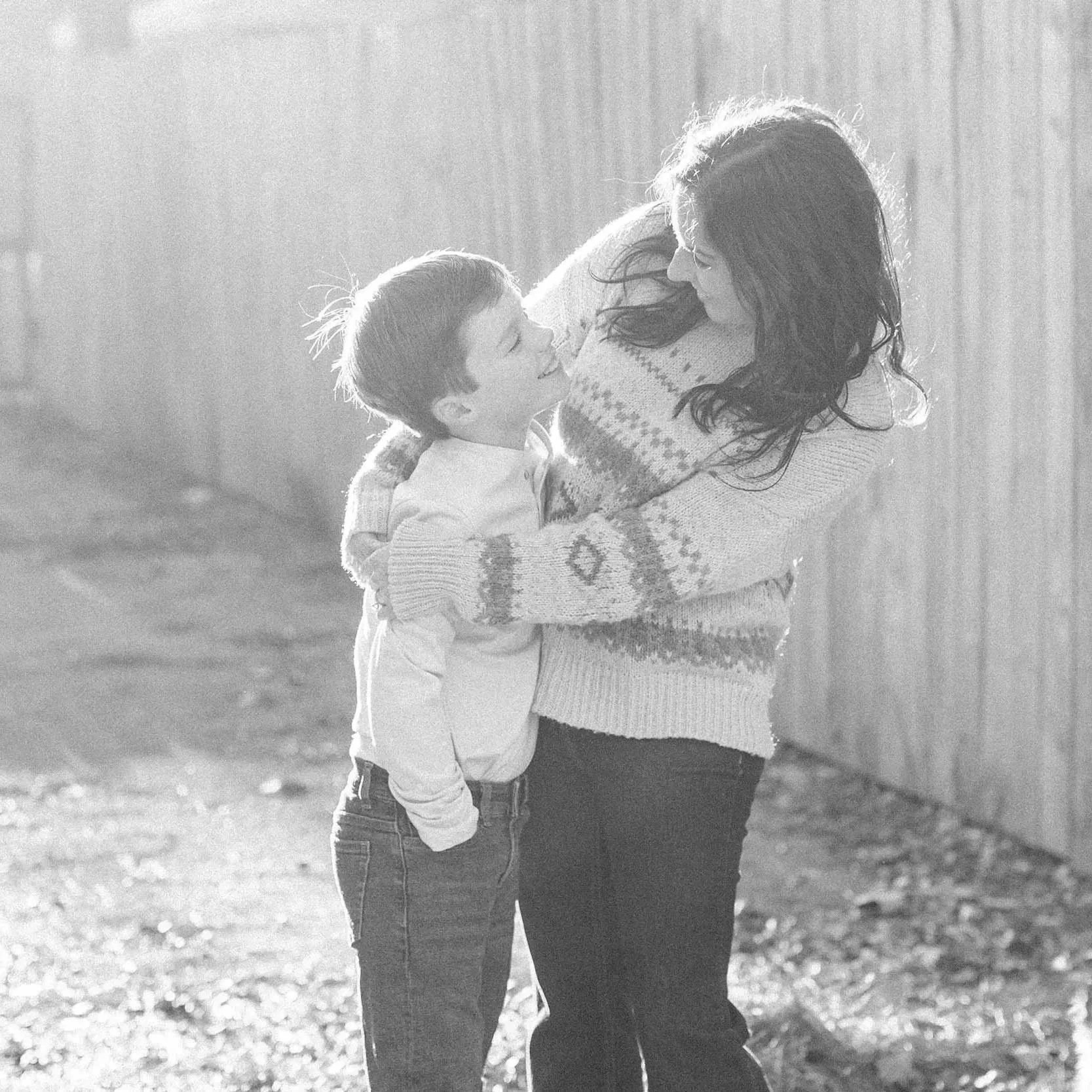 A woman with long hair holding a young boy in her arms outdoors, both smiling and facing each other in a tender moment, backlit with sunlight.