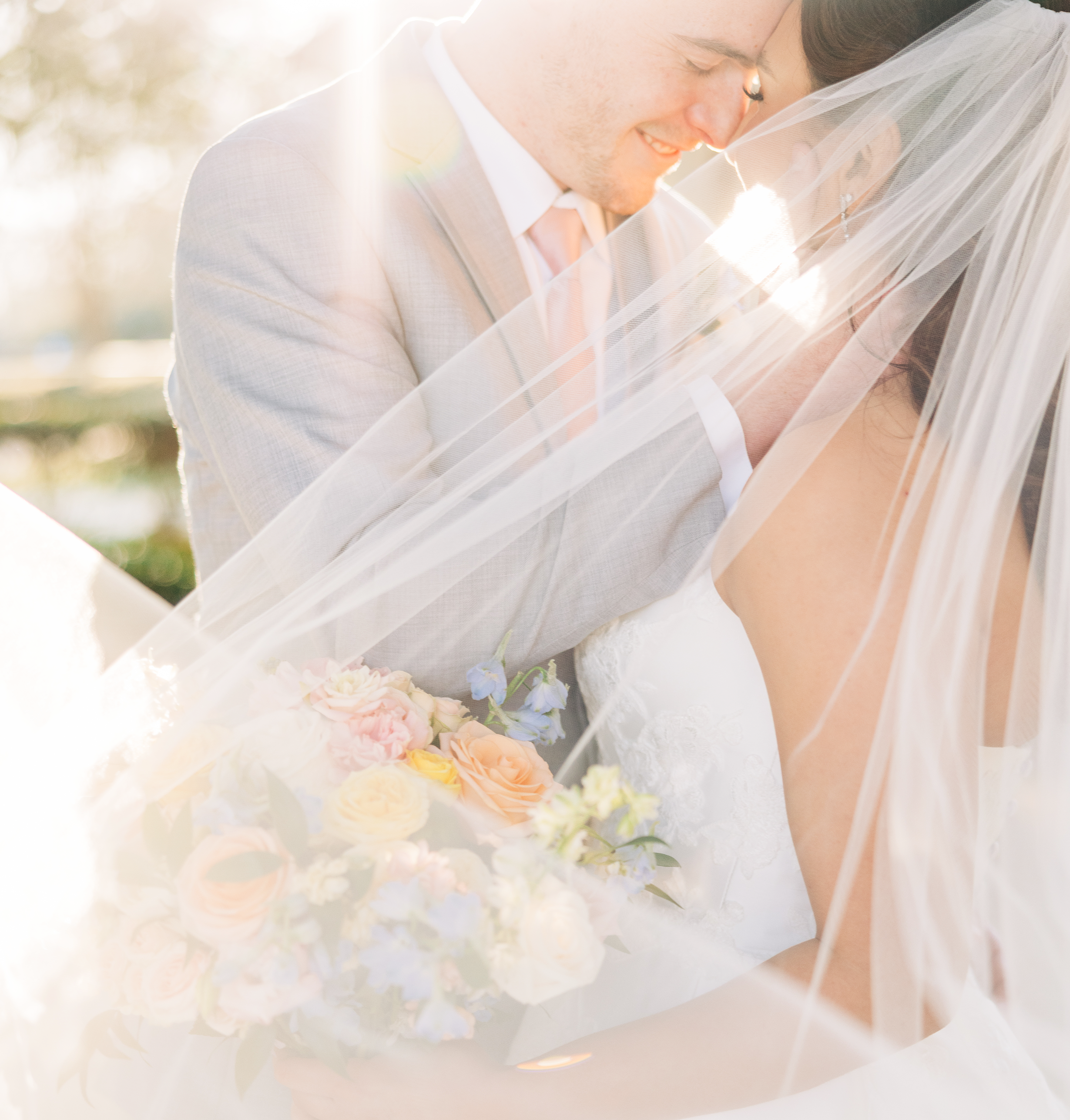 A close-up of a newlywed couple smiling and leaning their foreheads together, with the bride wearing a veil and holding a bouquet of pastel-colored flowers, backlit by soft sunlight.