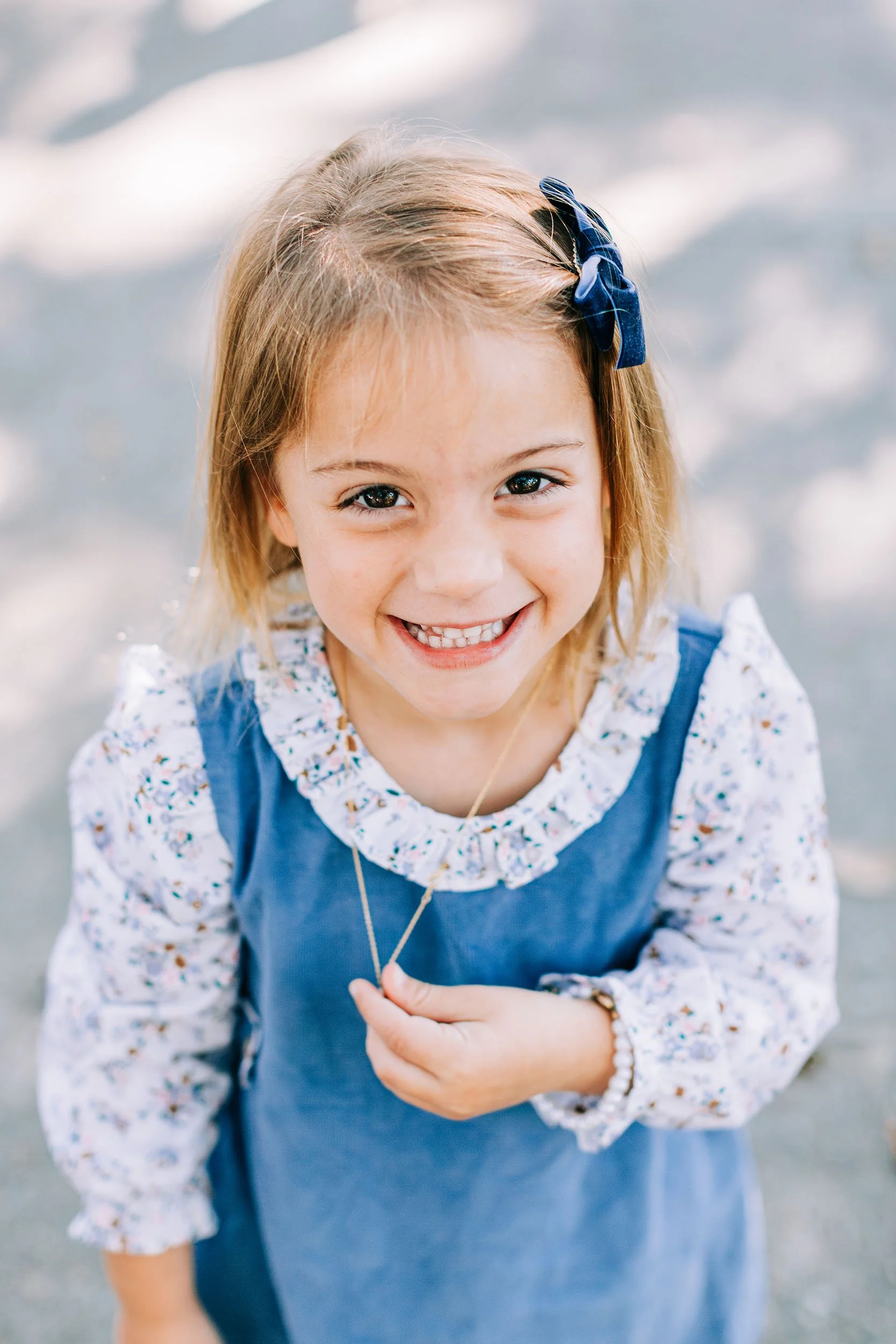 Close-up of a young girl with blonde hair tied with a blue ribbon, smiling at the camera, wearing a floral shirt and blue dress, standing at the beach with water and waves in the background.