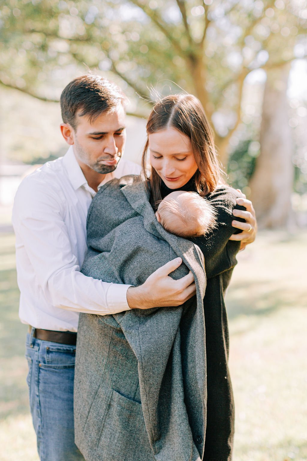 mom and dad holding and looking down at newborn baby wrapped in dad’s jacket during family portrait session in baton rouge