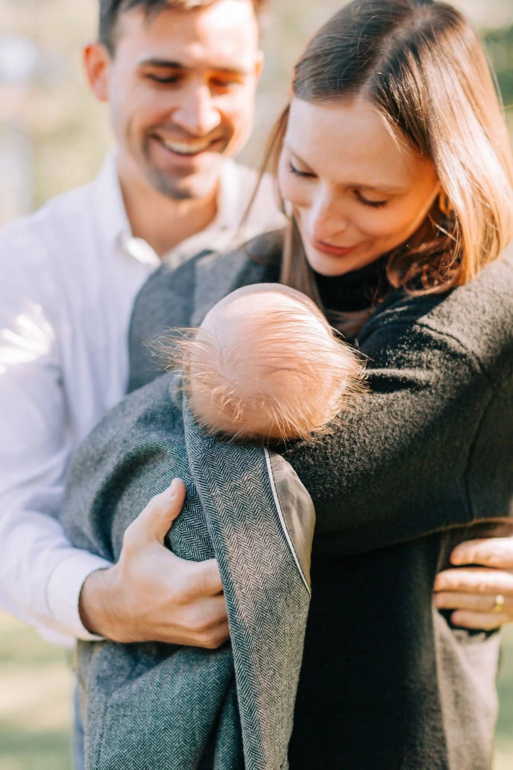 mom and dad holding and looking down at newborn baby wrapped in dad’s jacket during family portrait session in baton rouge