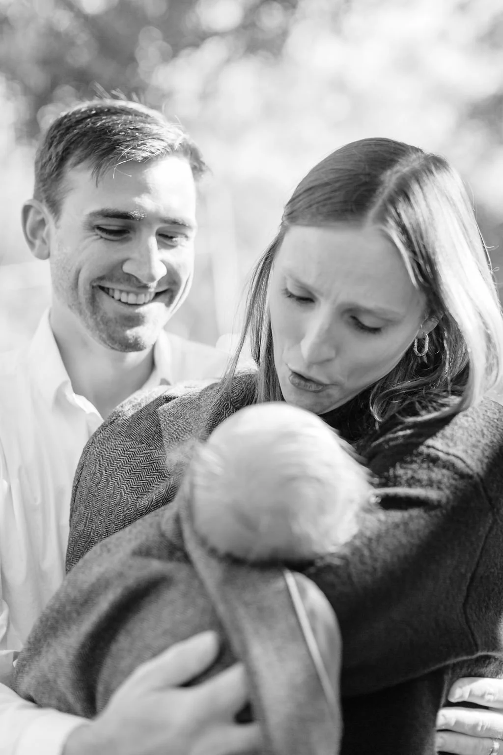 black and white moment of mom and dad holding and looking down at newborn baby wrapped in dad’s jacket during family portrait session in baton rouge