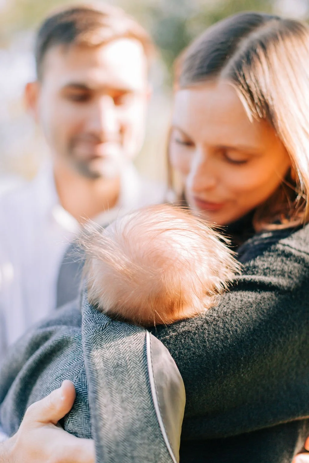 mom and dad holding and looking down at newborn baby wrapped in dad’s jacket during family portrait session in baton rouge