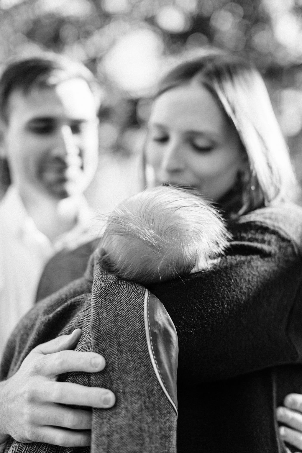 black and white moment of mom and dad holding and looking down at newborn baby wrapped in dad’s jacket, close-up of newborn baby’s hair during family portrait session in baton rouge