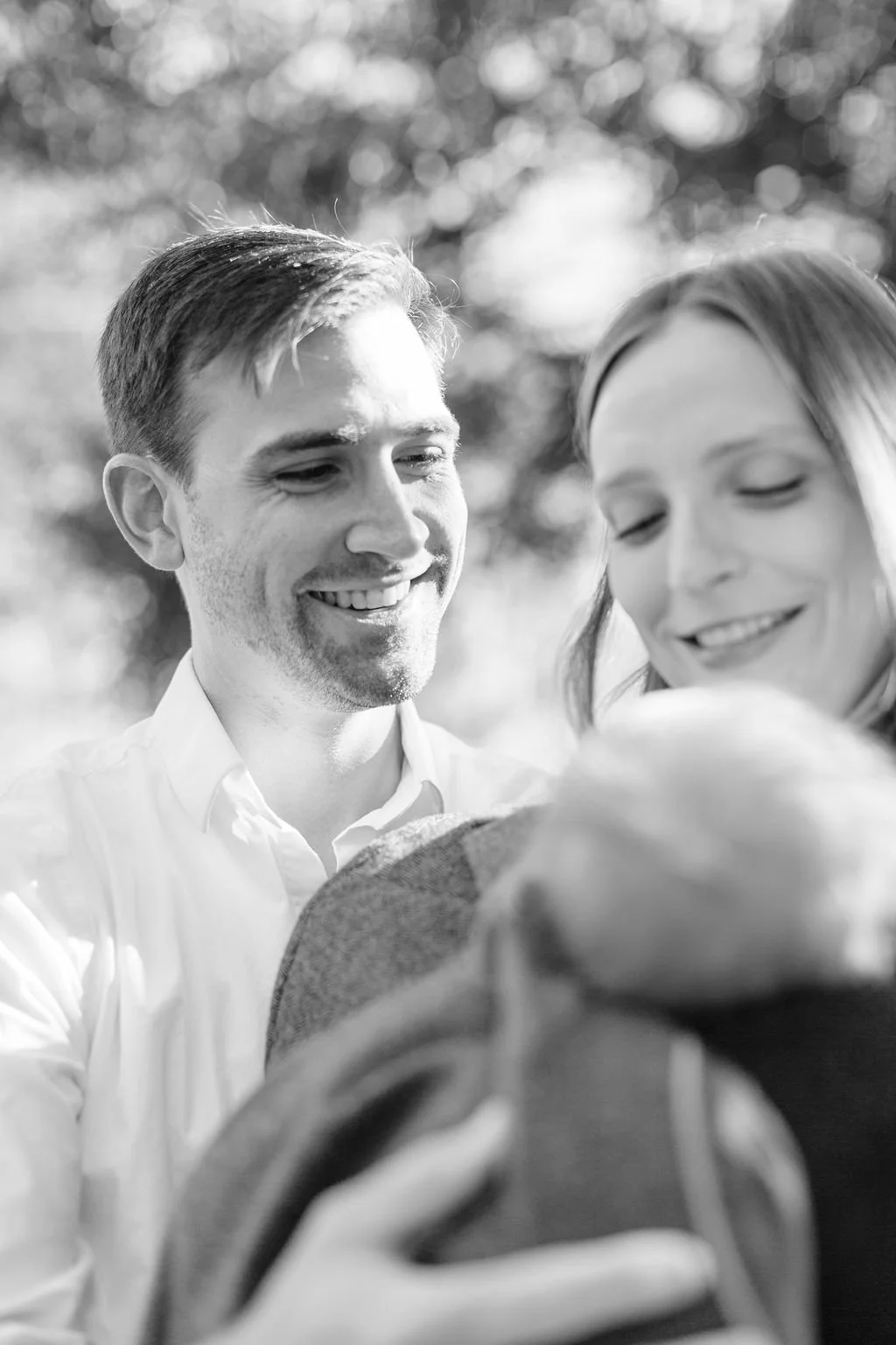 black and white moment of mom and dad holding and looking down at newborn baby wrapped in dad’s jacket, close-up of newborn baby’s hair during family portrait session in baton rouge
