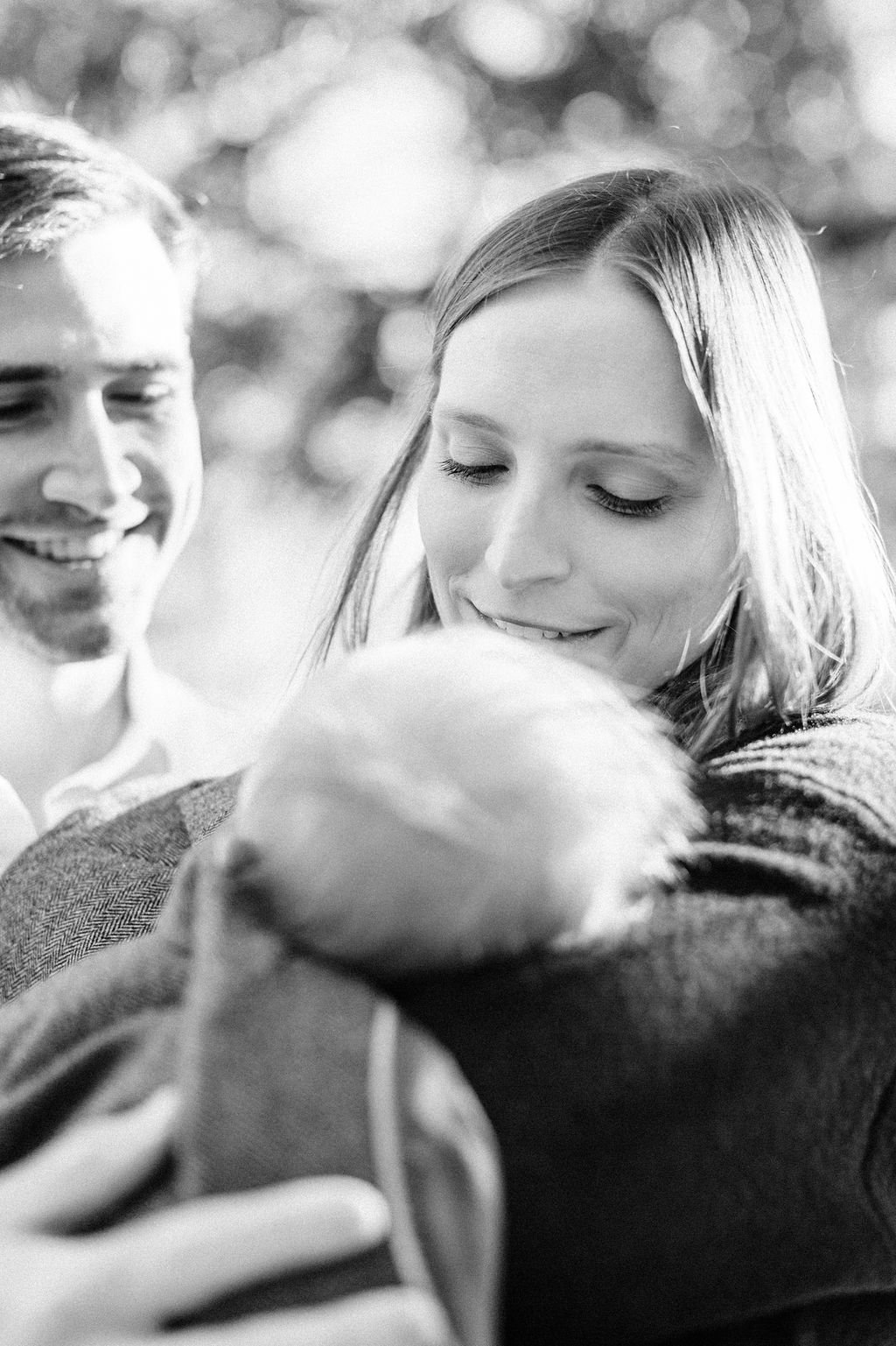 black and white moment of mom and dad holding and looking down at newborn baby wrapped in dad’s jacket, close-up of newborn baby’s hair during family portrait session in baton rouge