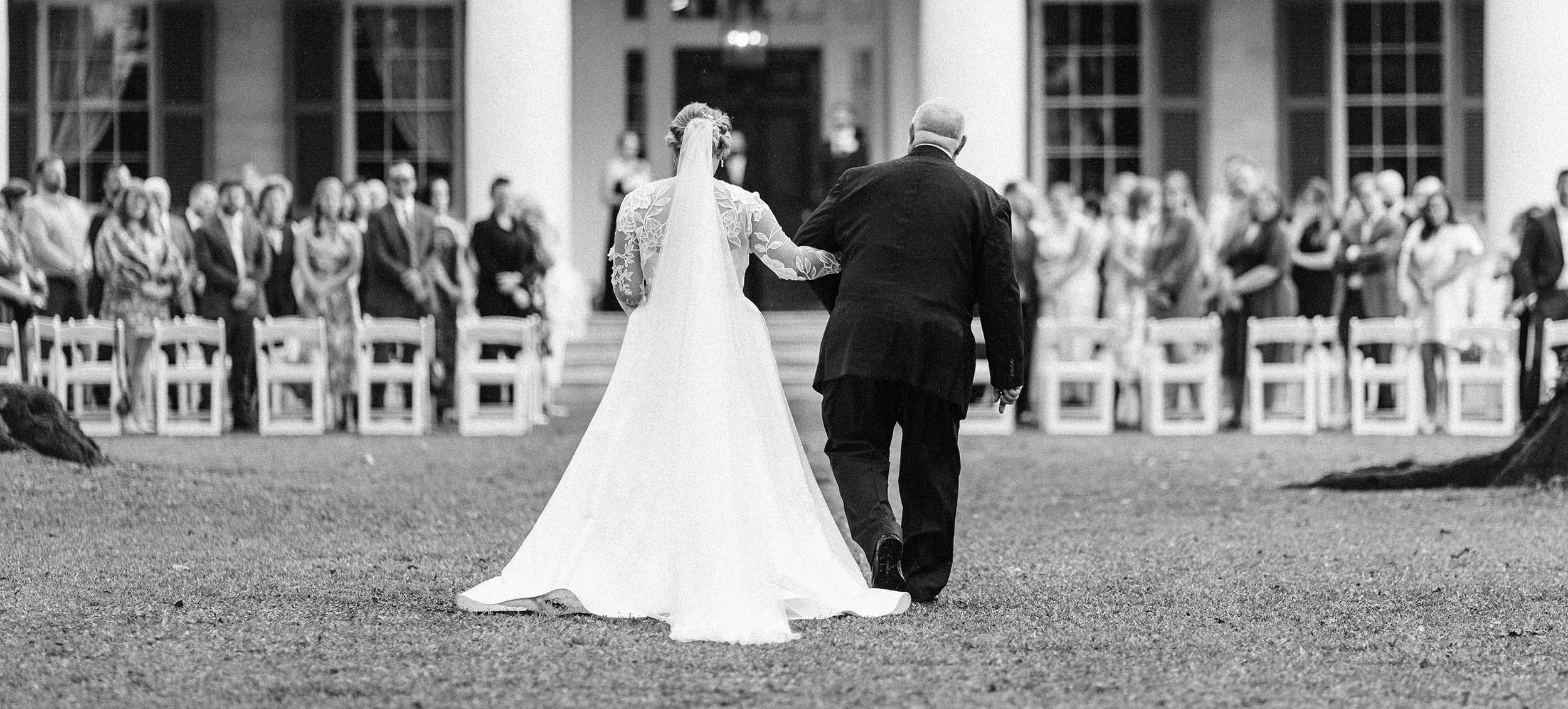 A bride walking down the aisle with her father at an outdoor wedding ceremony, with guests seated and standing as witnesses.