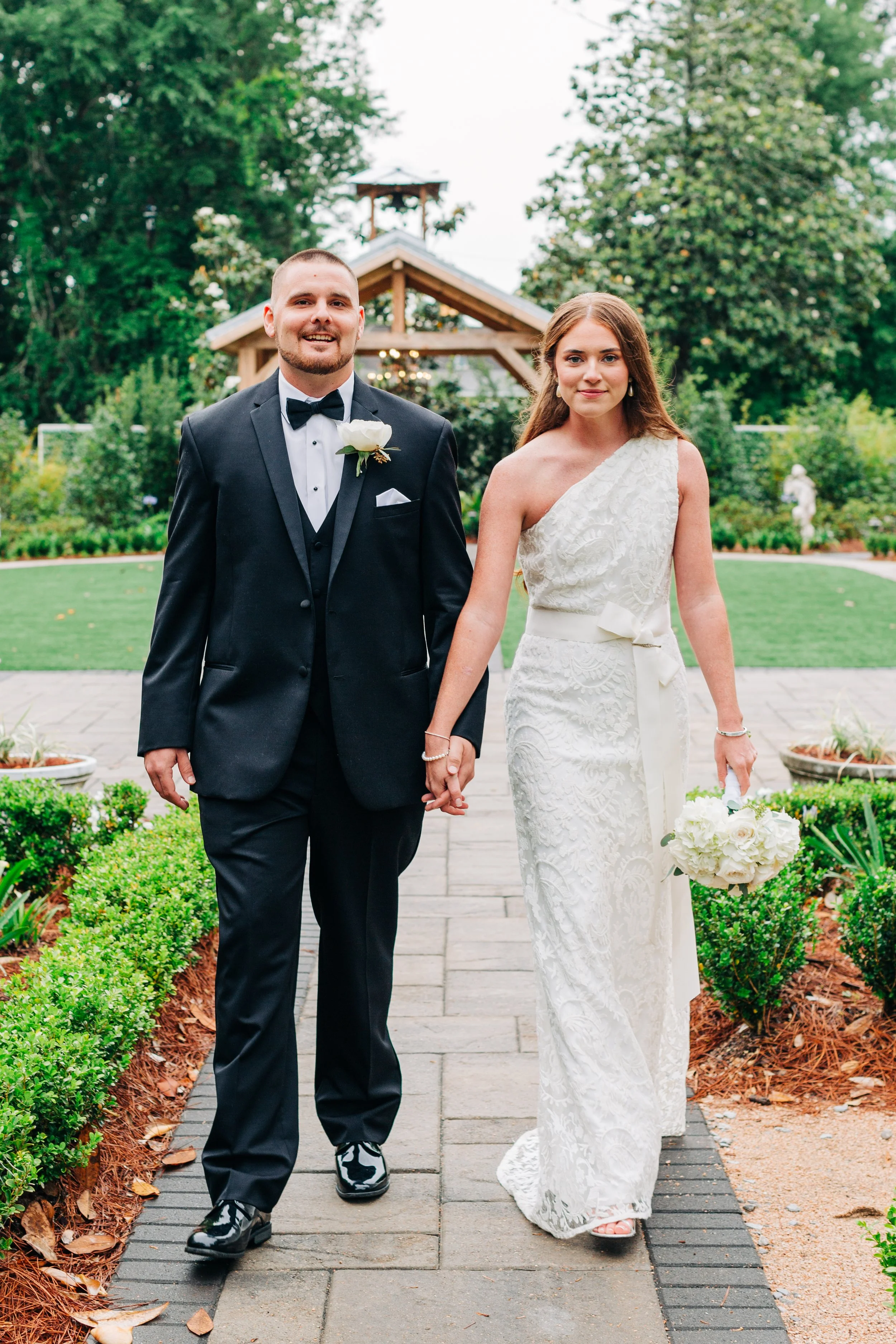 A bride and groom walking hand in hand outdoors during a wedding ceremony in a garden with lush greenery and statues, smiling at the camera.