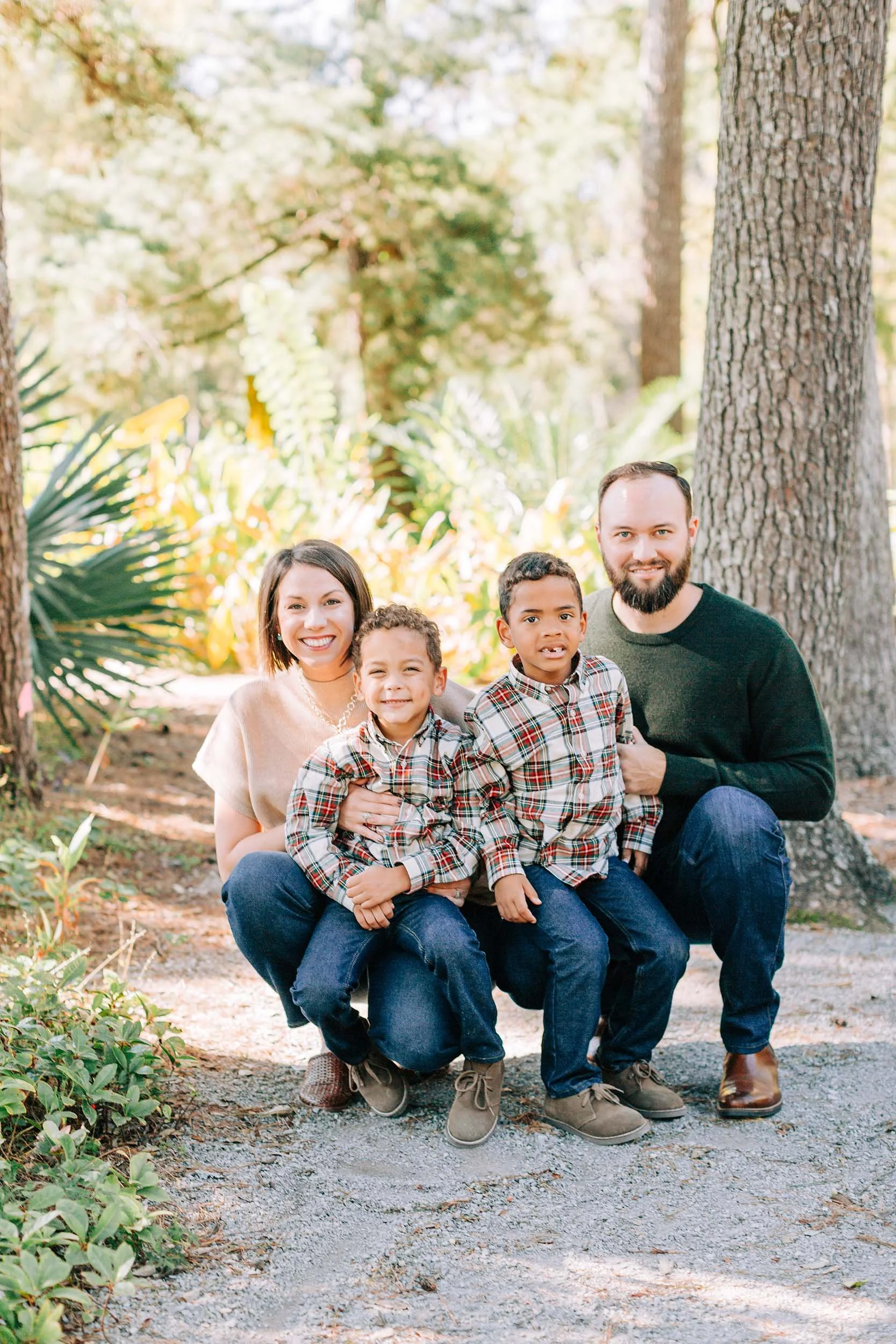 A family of four, including mother, father, and two young boys, posing outdoors in a wooded area with trees and green foliage in the background.