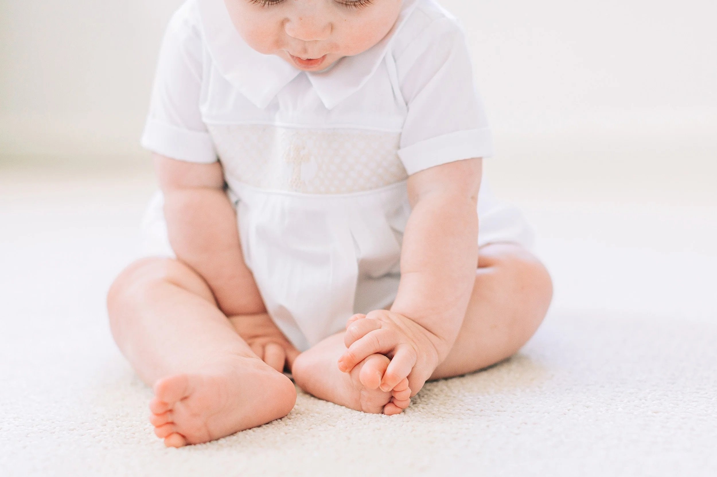 A young child sitting on a cream-colored carpet, wearing a white shirt and looking down with a focused expression.