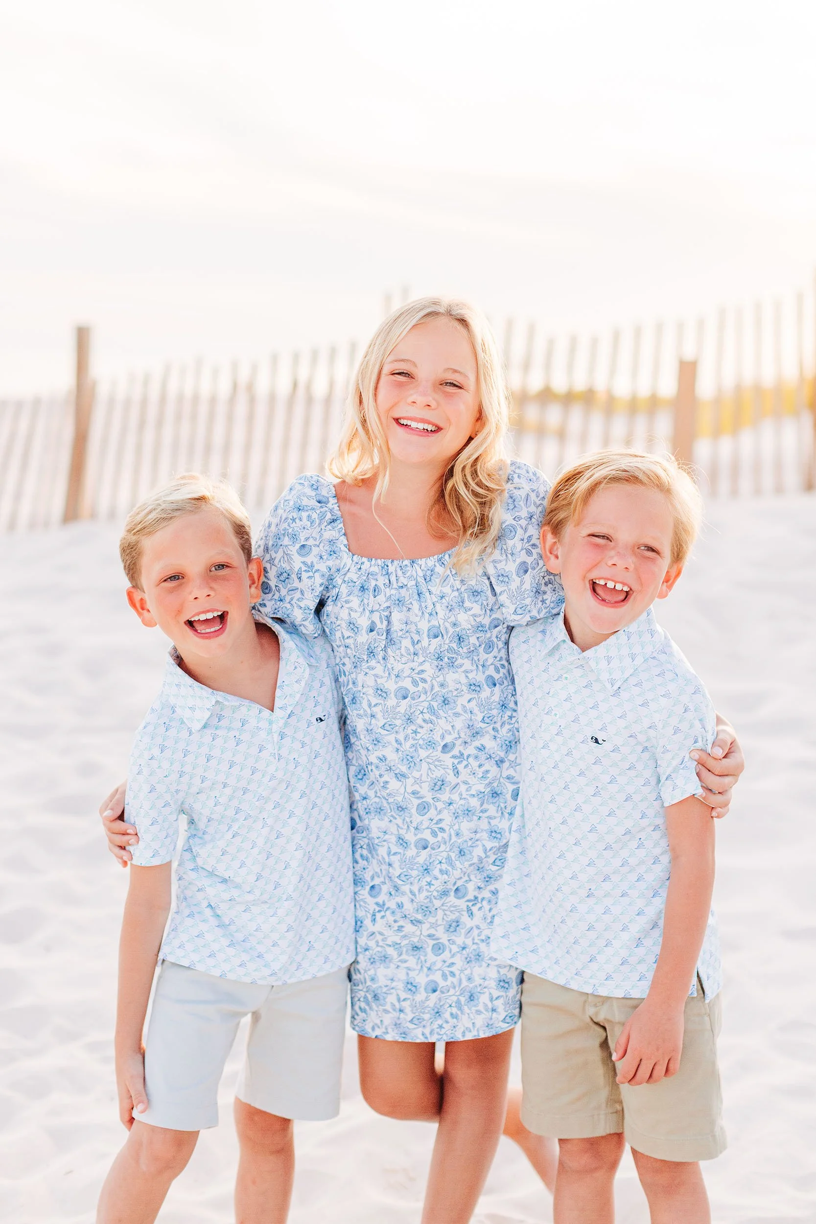A woman with long blonde hair and two young boys, all smiling, standing on a sandy beach with a wooden fence in the background.