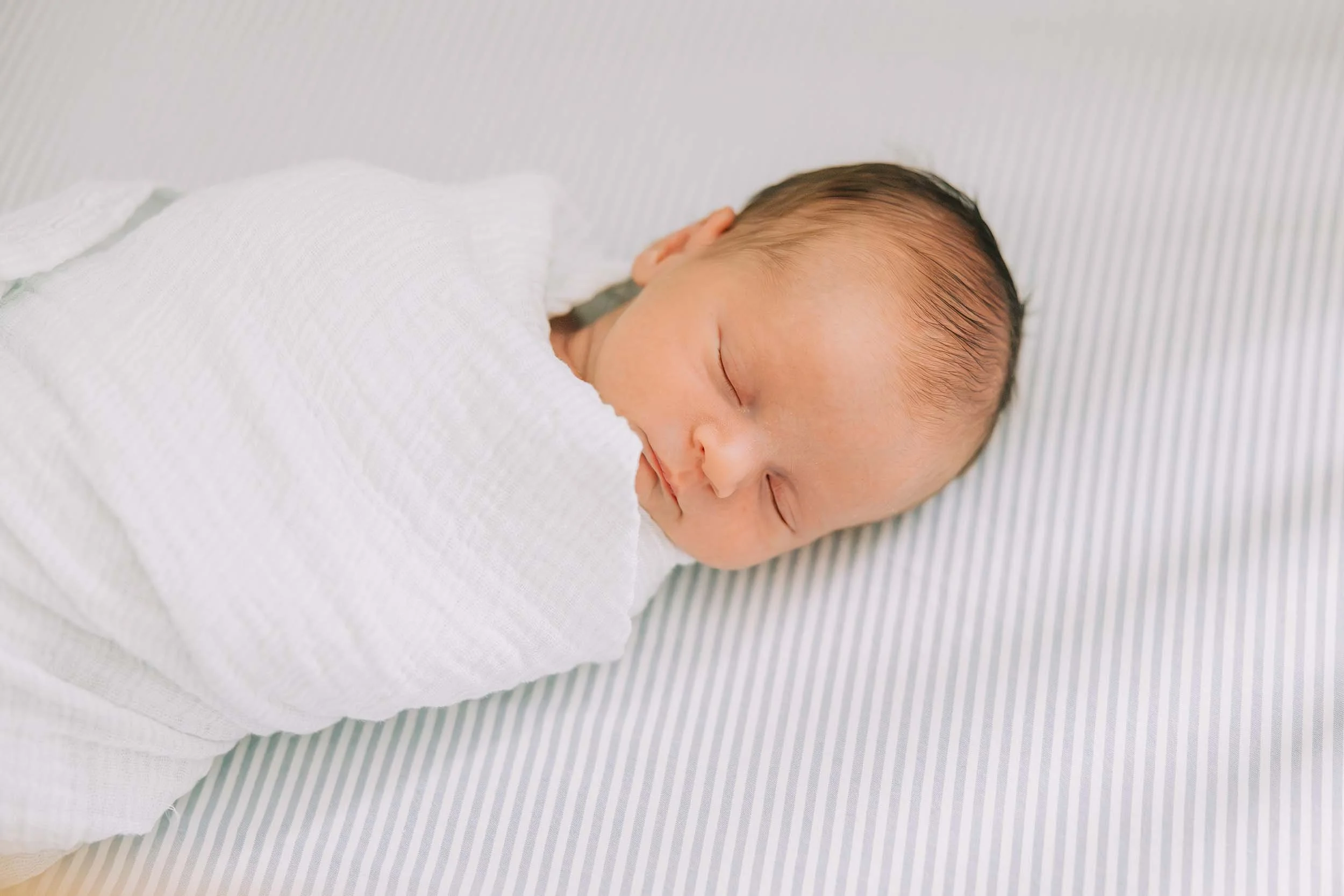 Close-up of a newborn baby sleeping on a striped surface, wrapped in a white blanket.
