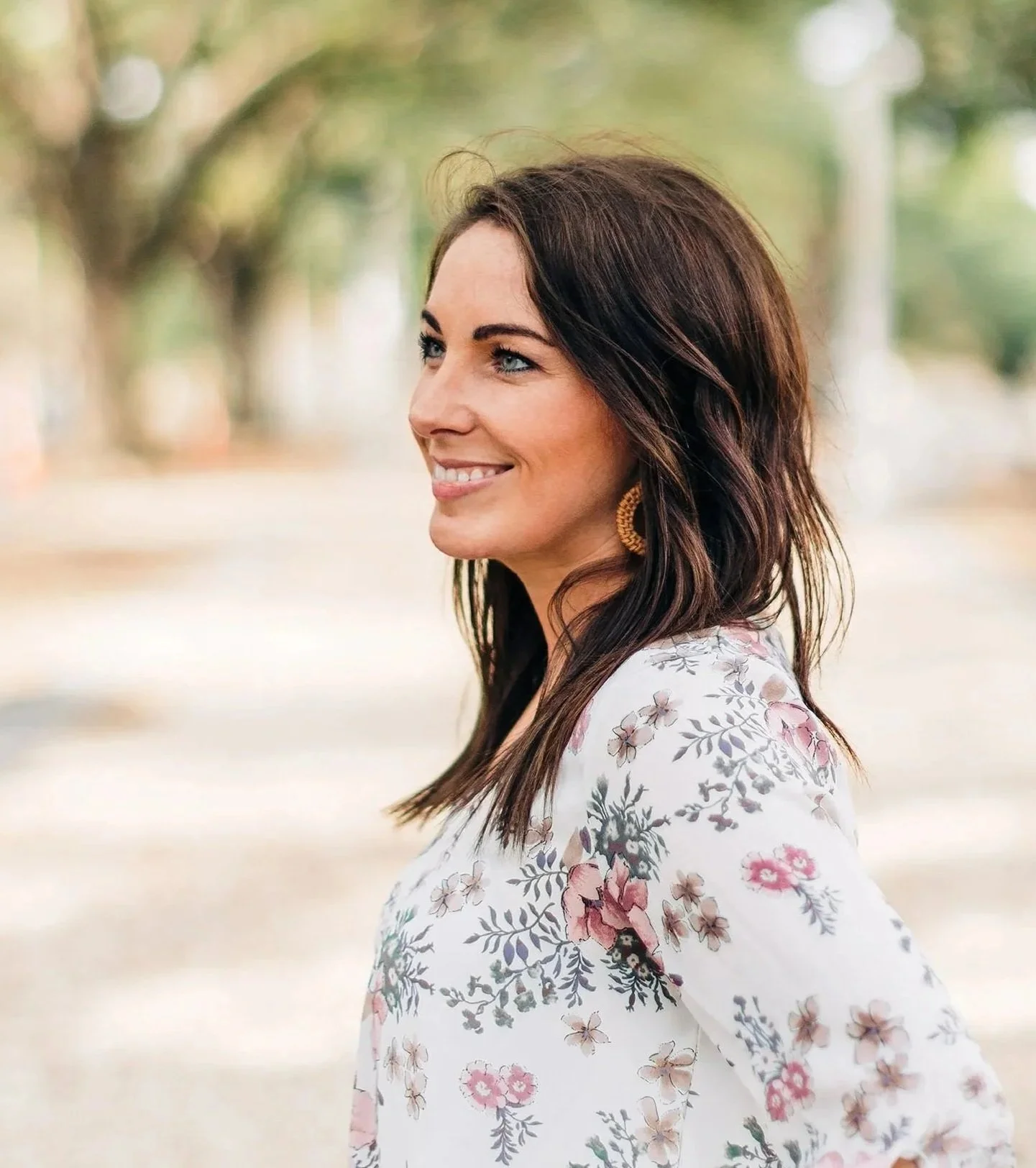 A woman with long dark hair wearing a floral patterned top and gold hoop earrings, smiling outdoors in a park with blurred trees in the background.