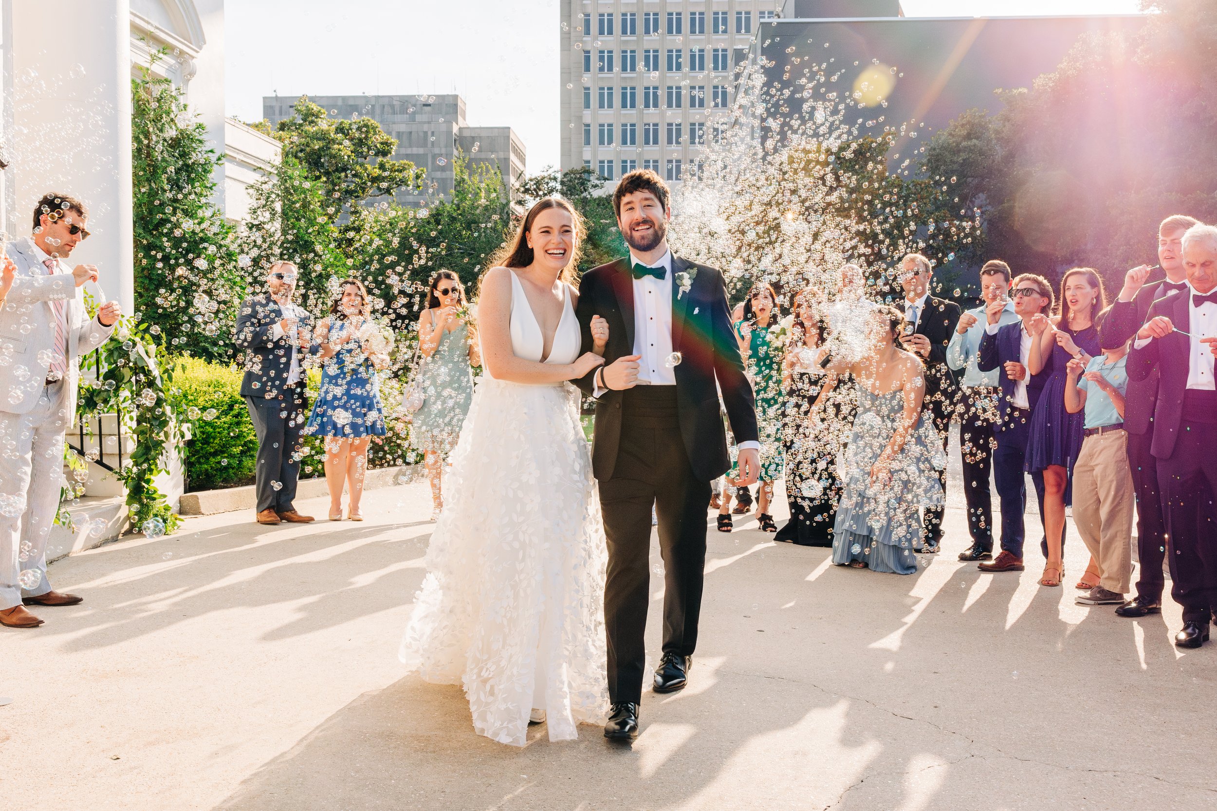 A bride and groom walking together at their outdoor wedding reception as guests celebrate and throw bubbles in the air, with city buildings in the background.