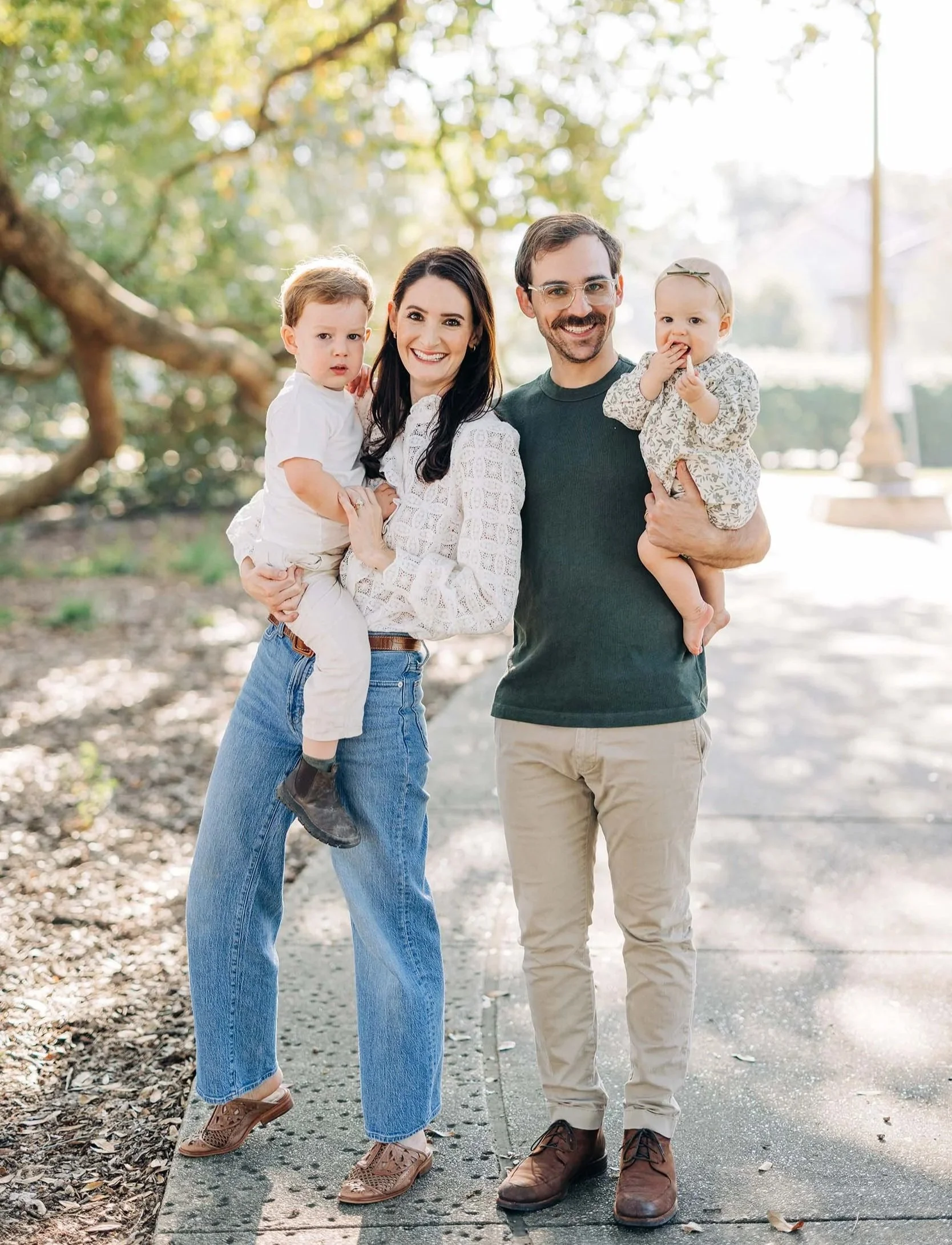 A happy family of four stands outdoors on a sunny day. The mother, with long dark hair, holds a young boy with light brown hair, while the father, with glasses and a beard, holds a young girl with a bow in her hair. They are surrounded by trees and natural light.