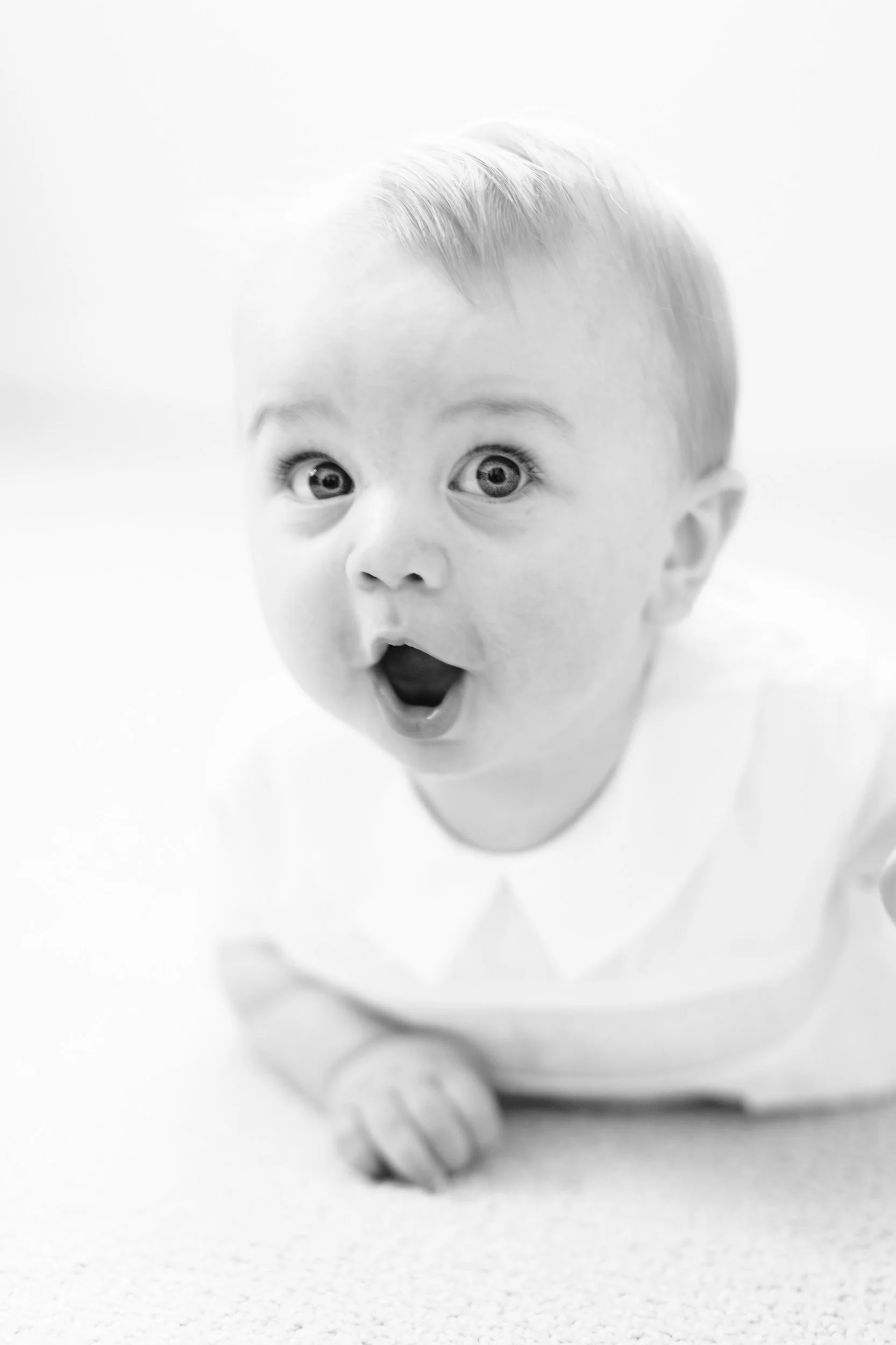 A black and white photo of a surprised baby crawling on a plain surface, with wide eyes and an open mouth.