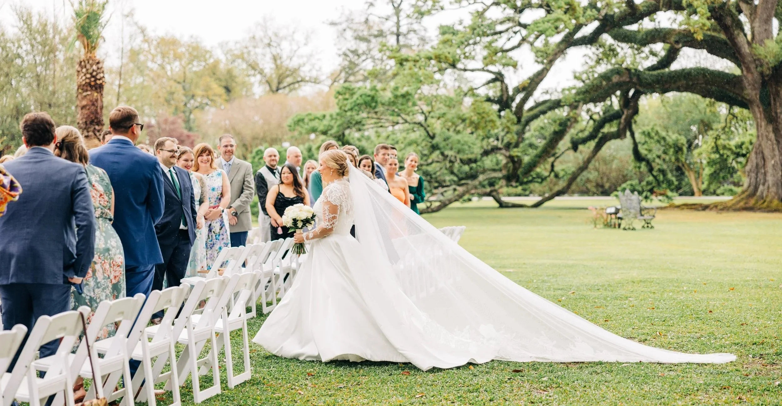 A bride in a wedding gown holding a bouquet, smiling as she walks down the aisle towards her guests during an outdoor wedding ceremony in a park with large trees.