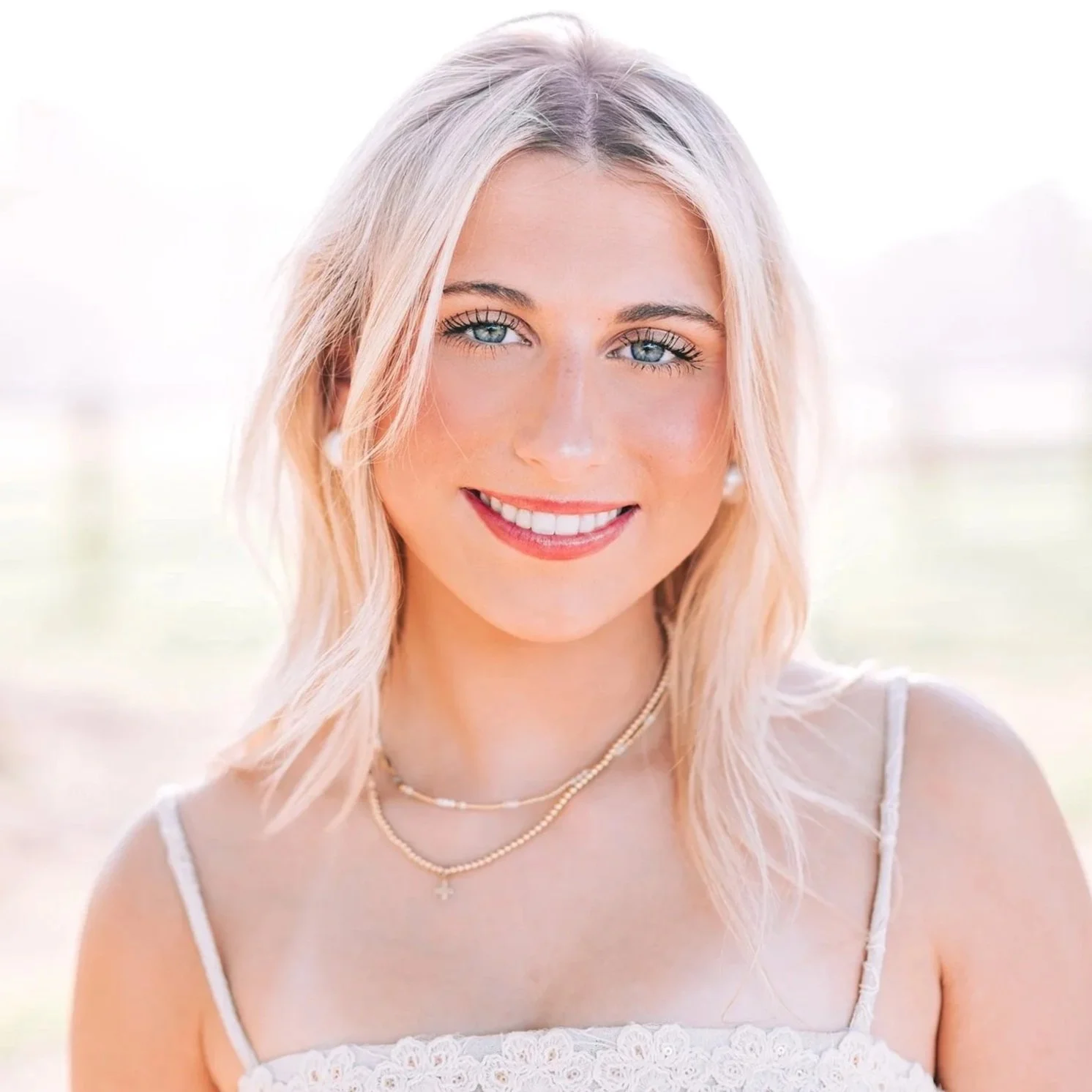 A young woman with blonde, wavy hair and blue eyes, smiling, wearing a white spaghetti strap top and layered necklaces, outdoors during daytime.