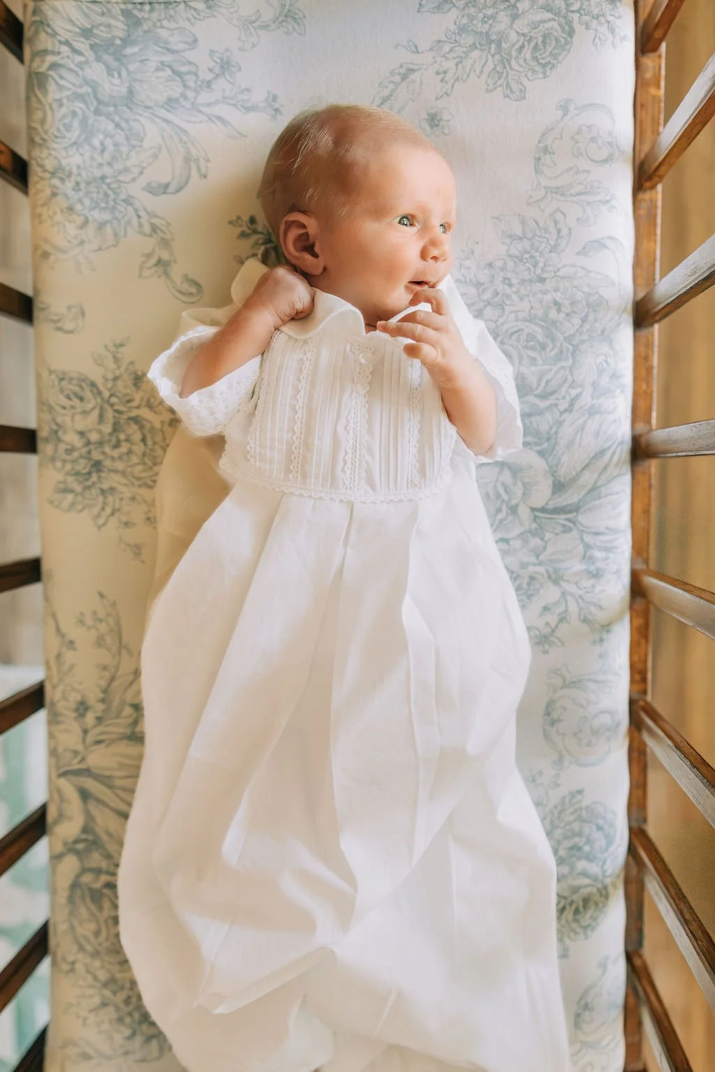 A baby in a white christening gown lying on a floral cushion on a wooden bed frame, gazing to the side with one finger near the mouth.