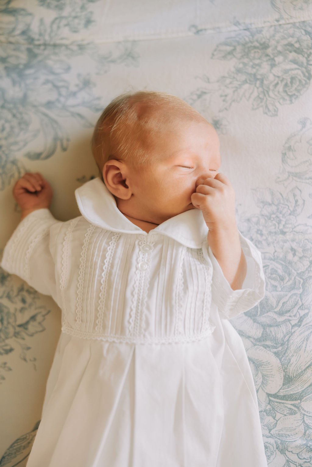 A newborn baby sleeping on a light-colored floral blanket, wearing a white embroidered gown, with hand near face and peaceful expression.