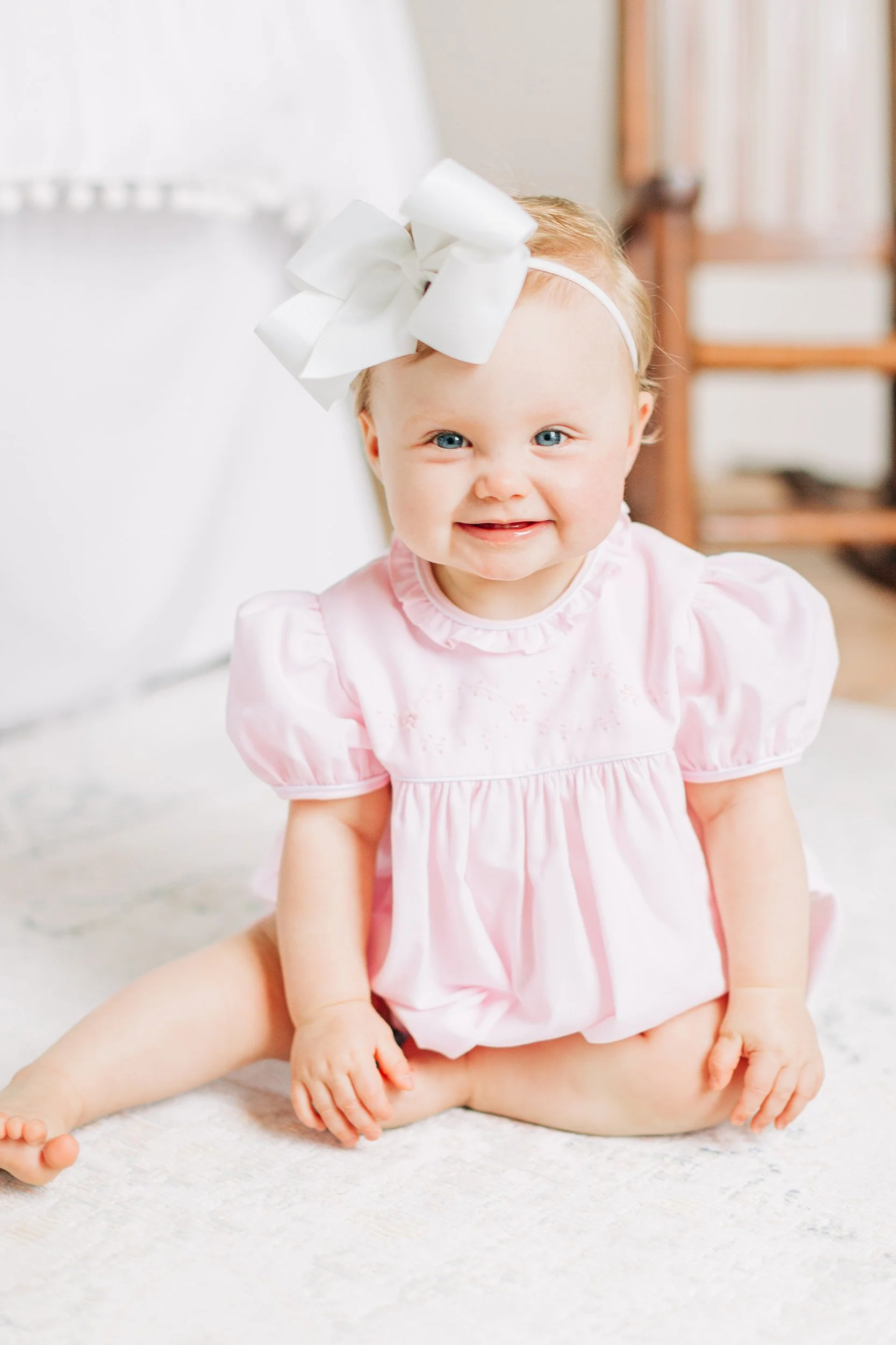 A smiling baby girl with blue eyes and red hair, wearing a pink dress and a large white bow headband, sitting on a white carpet in a room with wooden furniture and curtains.