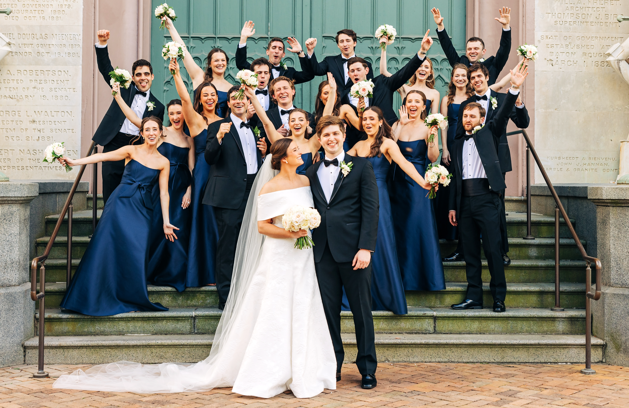 Large wedding party celebrating on steps with bride and groom surrounded by bridesmaids and groomsmen in Baton Rouge