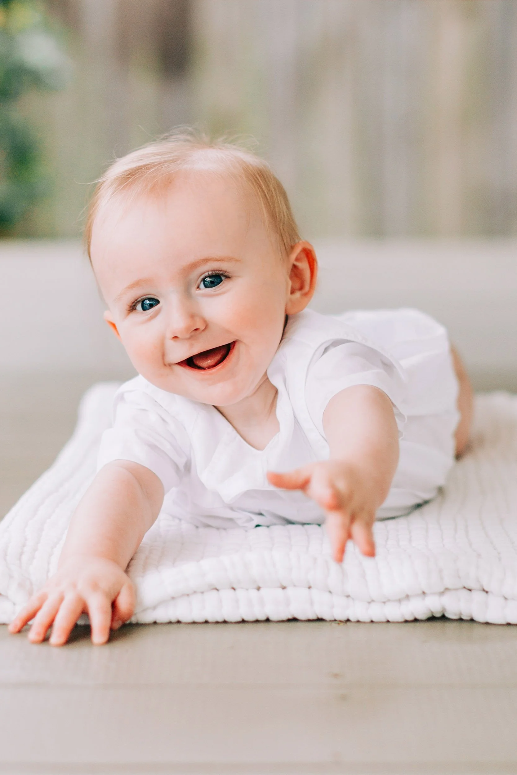 Close-up of a smiling baby with blue eyes reaching forward on a white blanket.